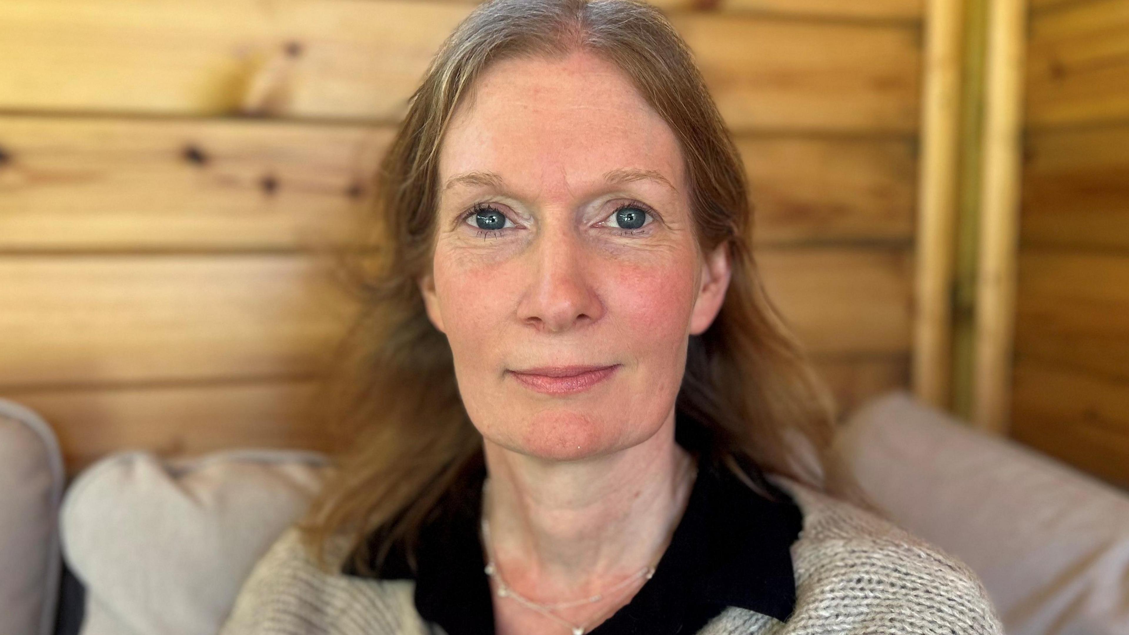 Melissa Robinson looking into the camera. She has brown hair which falls just below her shoulders. She is sitting on a grey sofa and in front of a wooden wall. She is wearing a black shirt with a cream jumper over the top and a silver necklace.