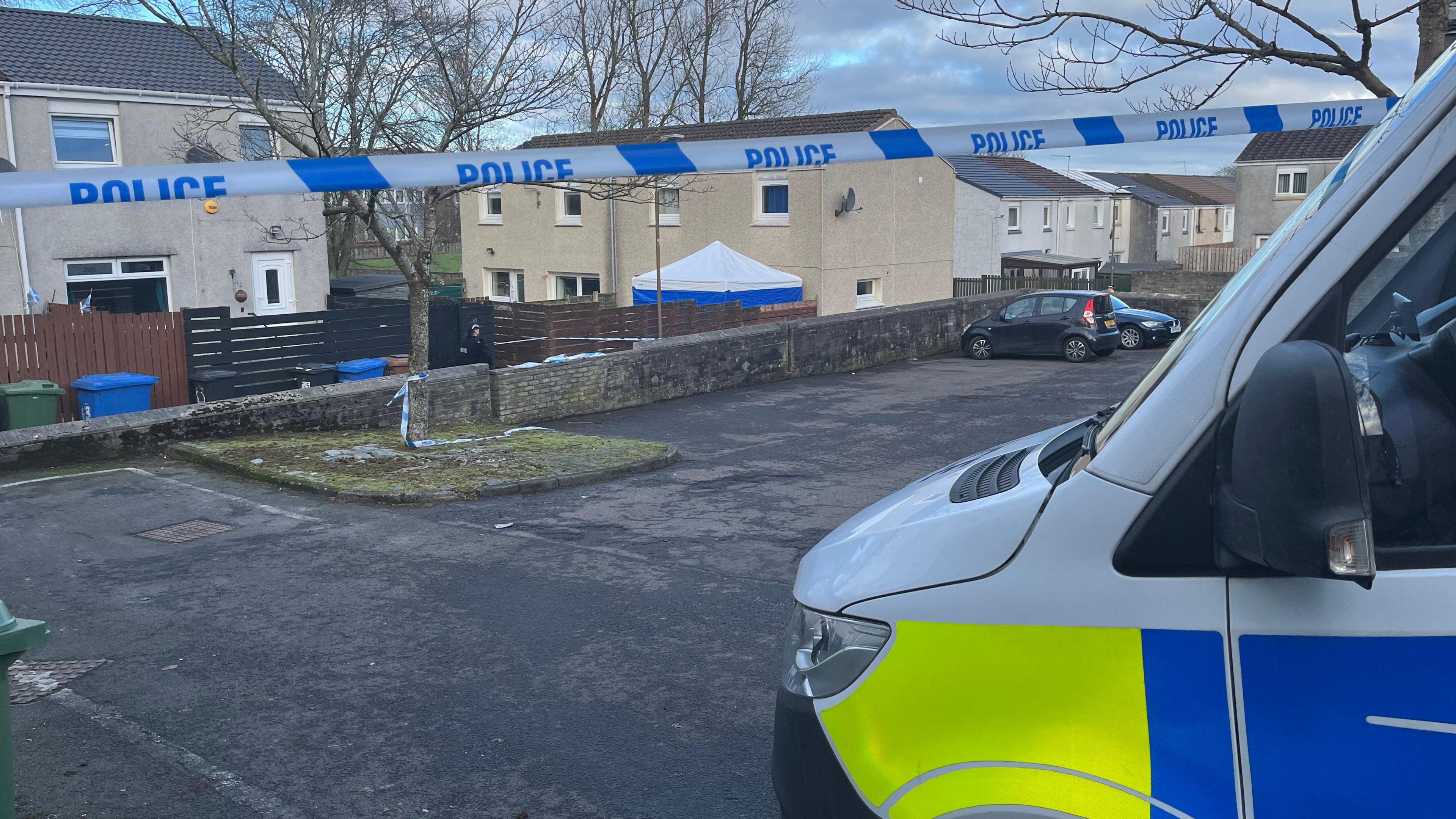 A view of Harburn Drive in West Calder. The house has a blue police forensic tent outside with a white roof. A police van is parked in the foreground in front of blue and white police tape.