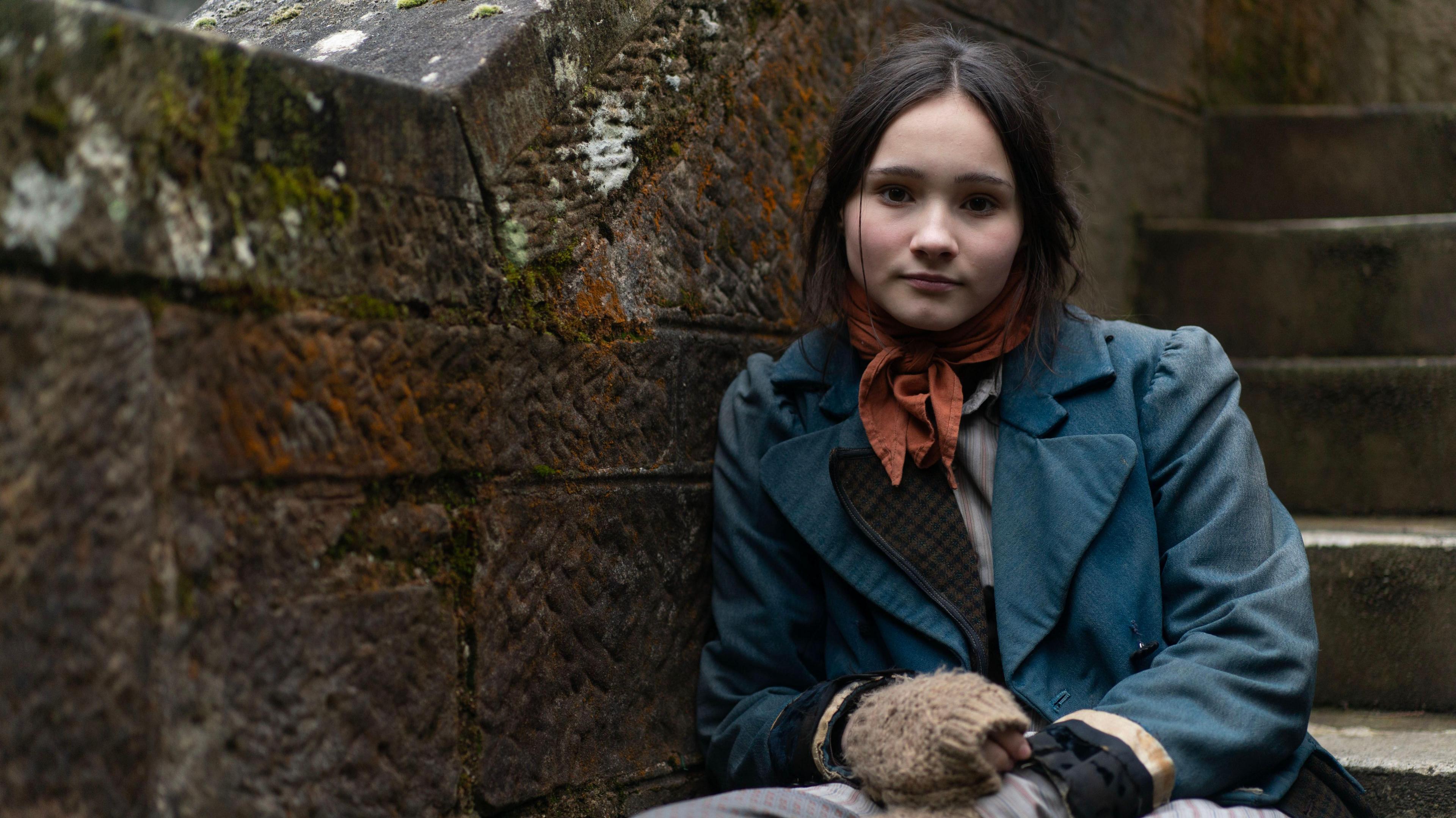 A young girl, sits on some steps outside next to a brick wall, She has dark hair which is tied back. She is wearing a period costume which is made up of a blue jacket, orange neck scarf and gloves.