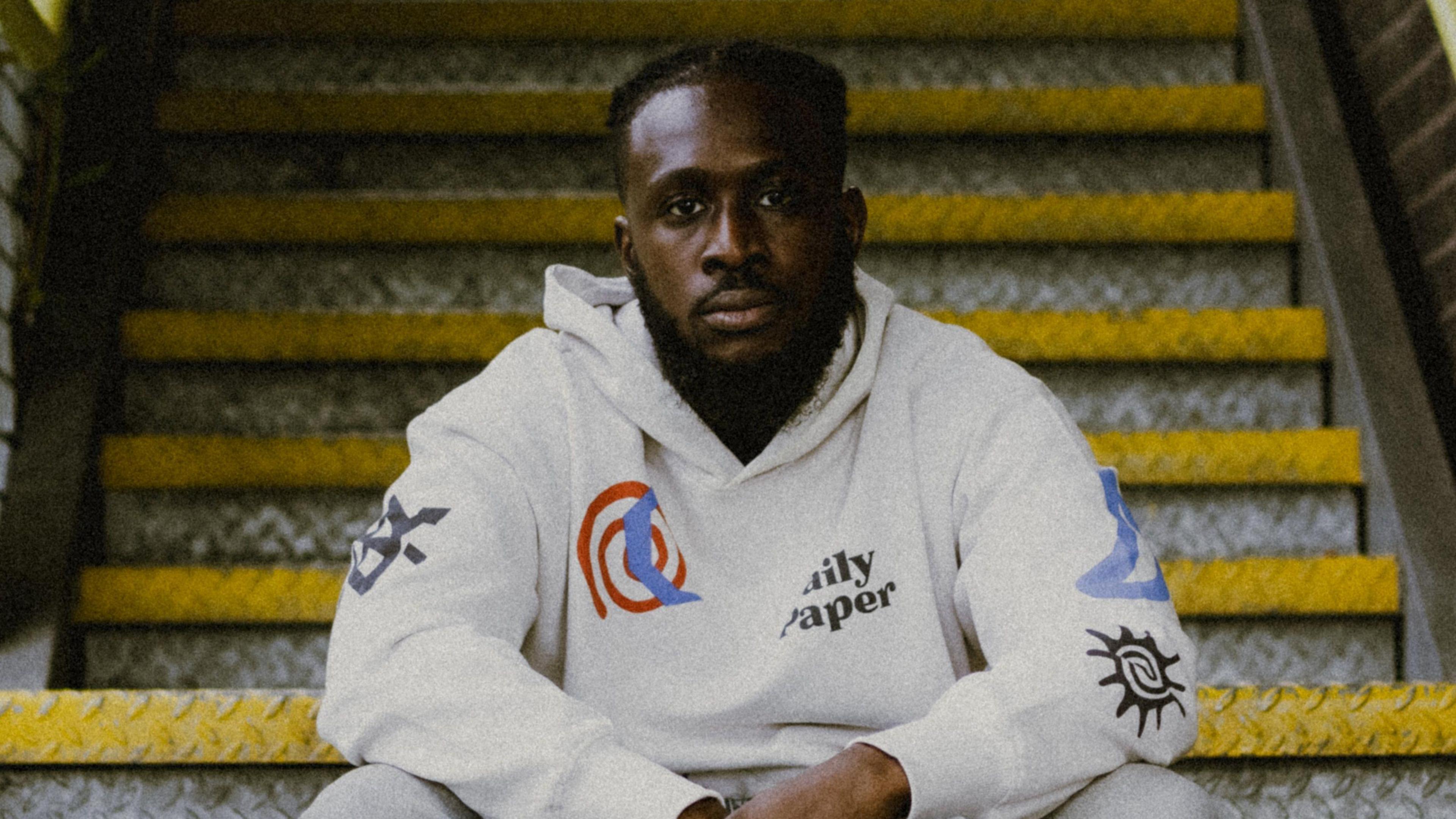 Still Shadey sits on some steel stairs outside, his arms folded in his lap. He has a trimmed beard, has his hair tied back wears a grey hoody while looking seriously at the camera. 