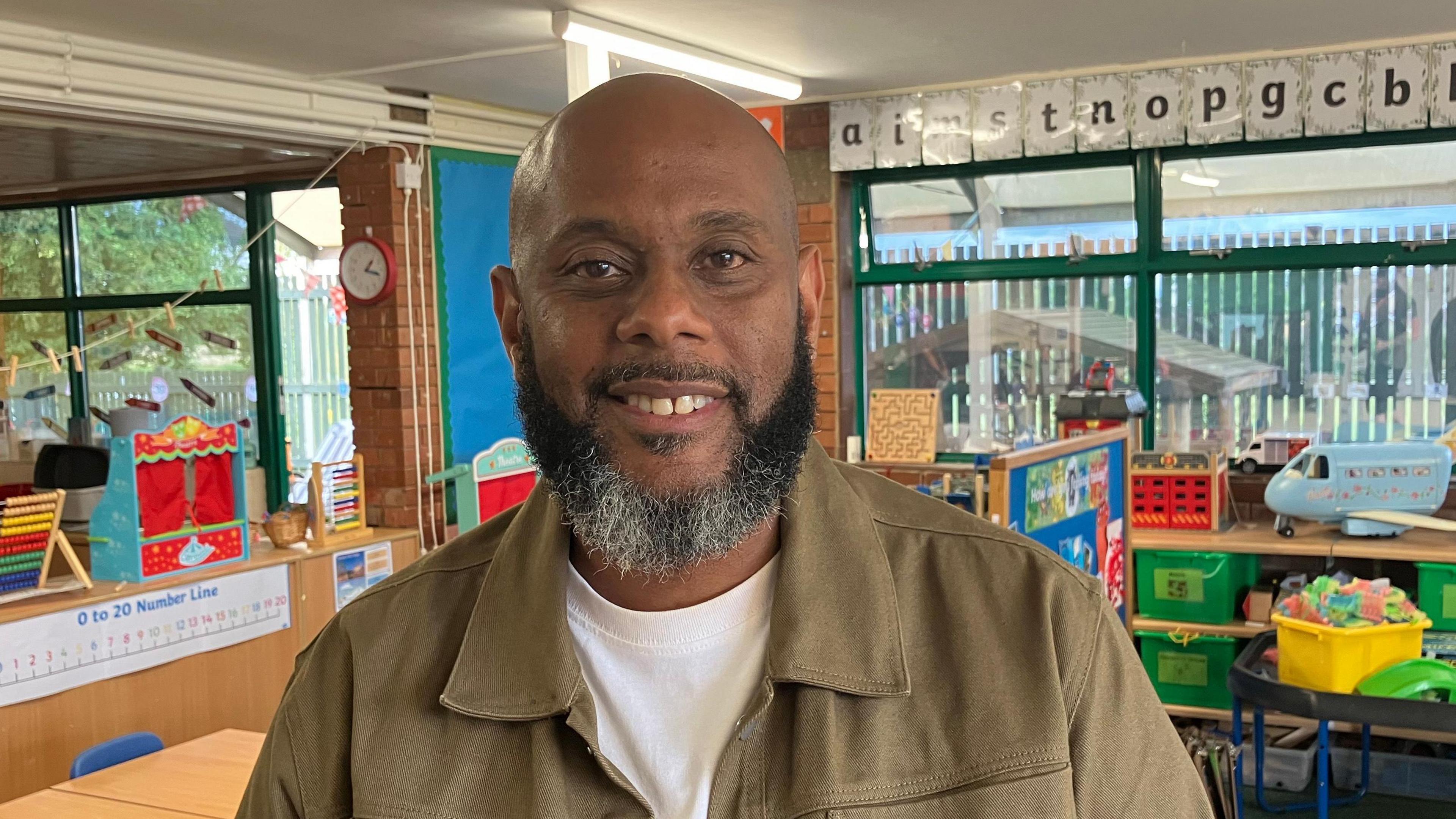 Gareth Fuller in a khaki coloured jacket and white T-shirt stands in a classroom.