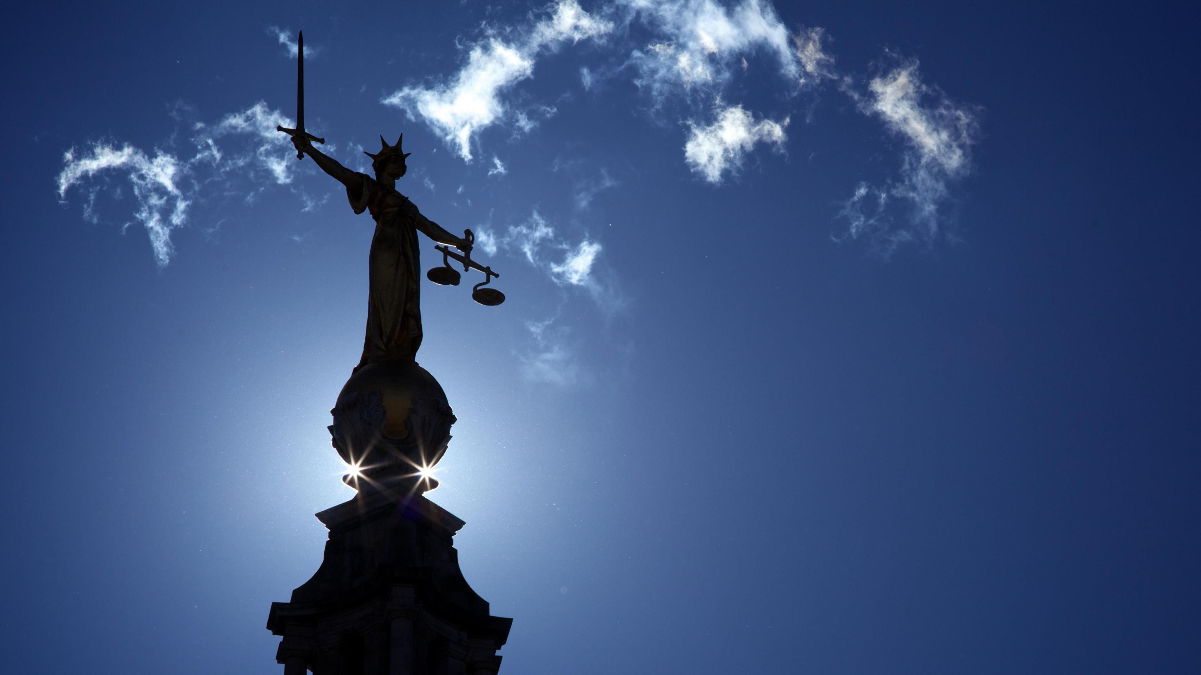 The scales of justice statue above the Old Bailey. The sun rises behind it. There is a blue sky and a few clouds.
