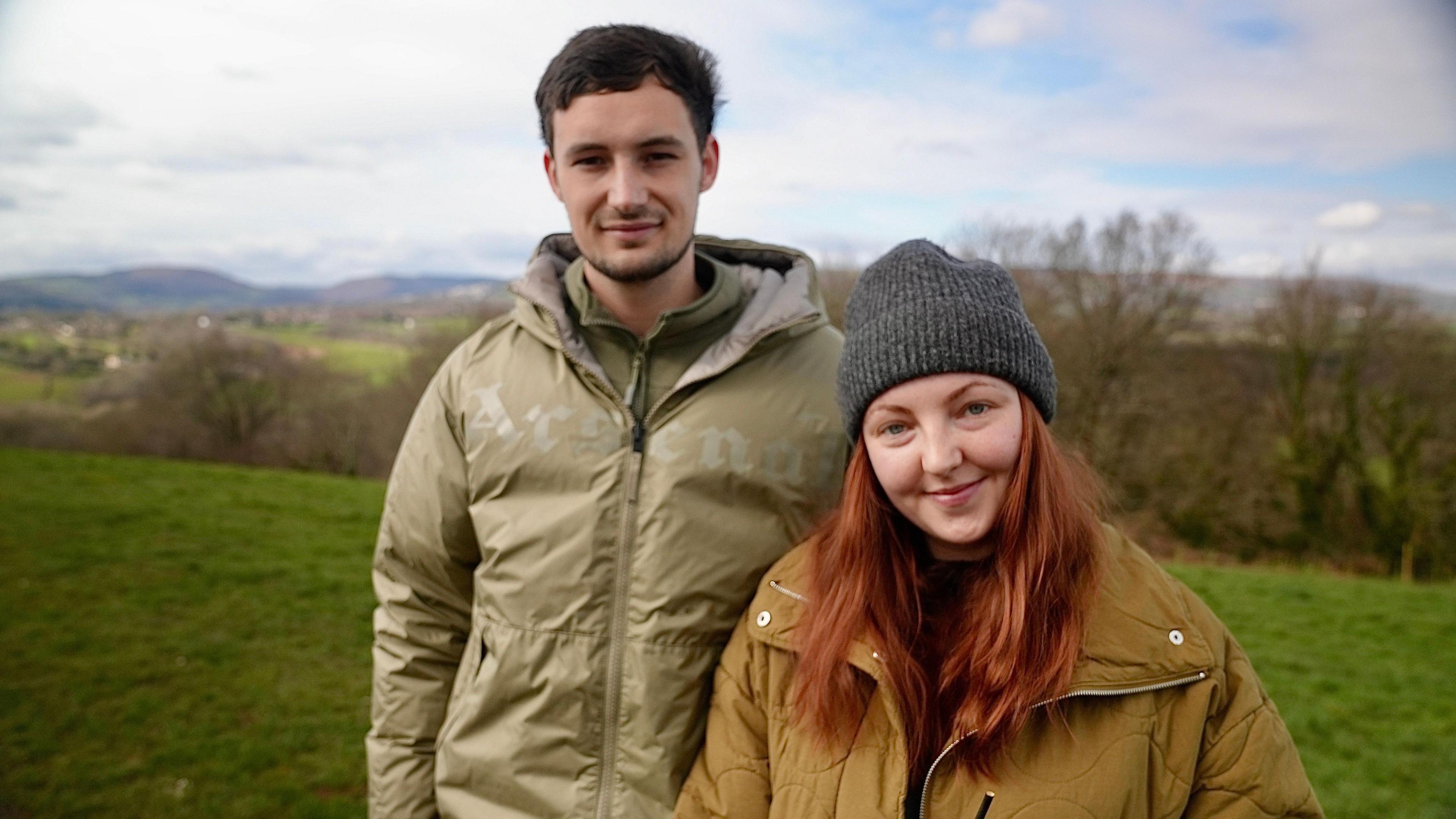 Jade and Jamie are standing in a meadow, side by side, Jade is wearing a beanie hat and Jamie a green coat.