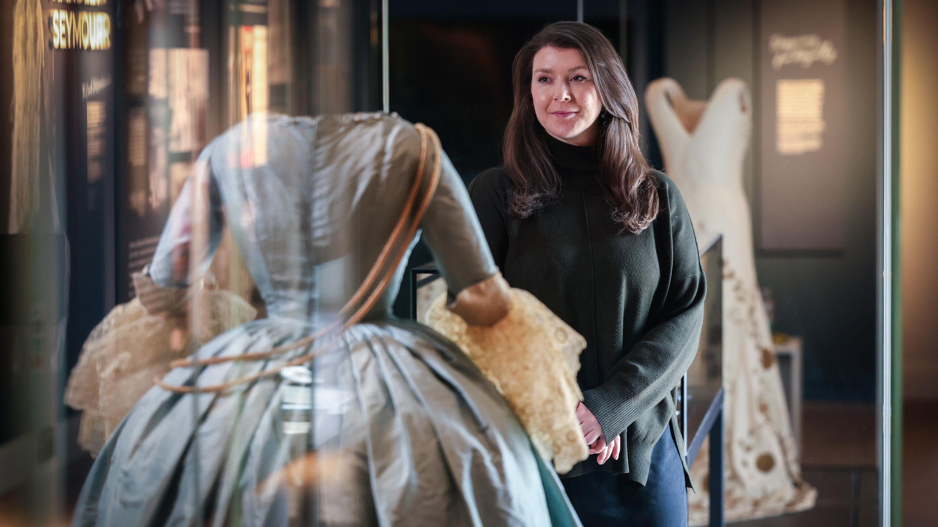 Claudia Acott Williams looking at one of the dresses in a glass cabinet. She has long brown hair, wearing a dark green jumper with her hands folded together. The dress is a Georgian-style costume gown in blue with white lace on the sleeves. The coronation toile can be seen in background. It is a white gown with gold embroidery.