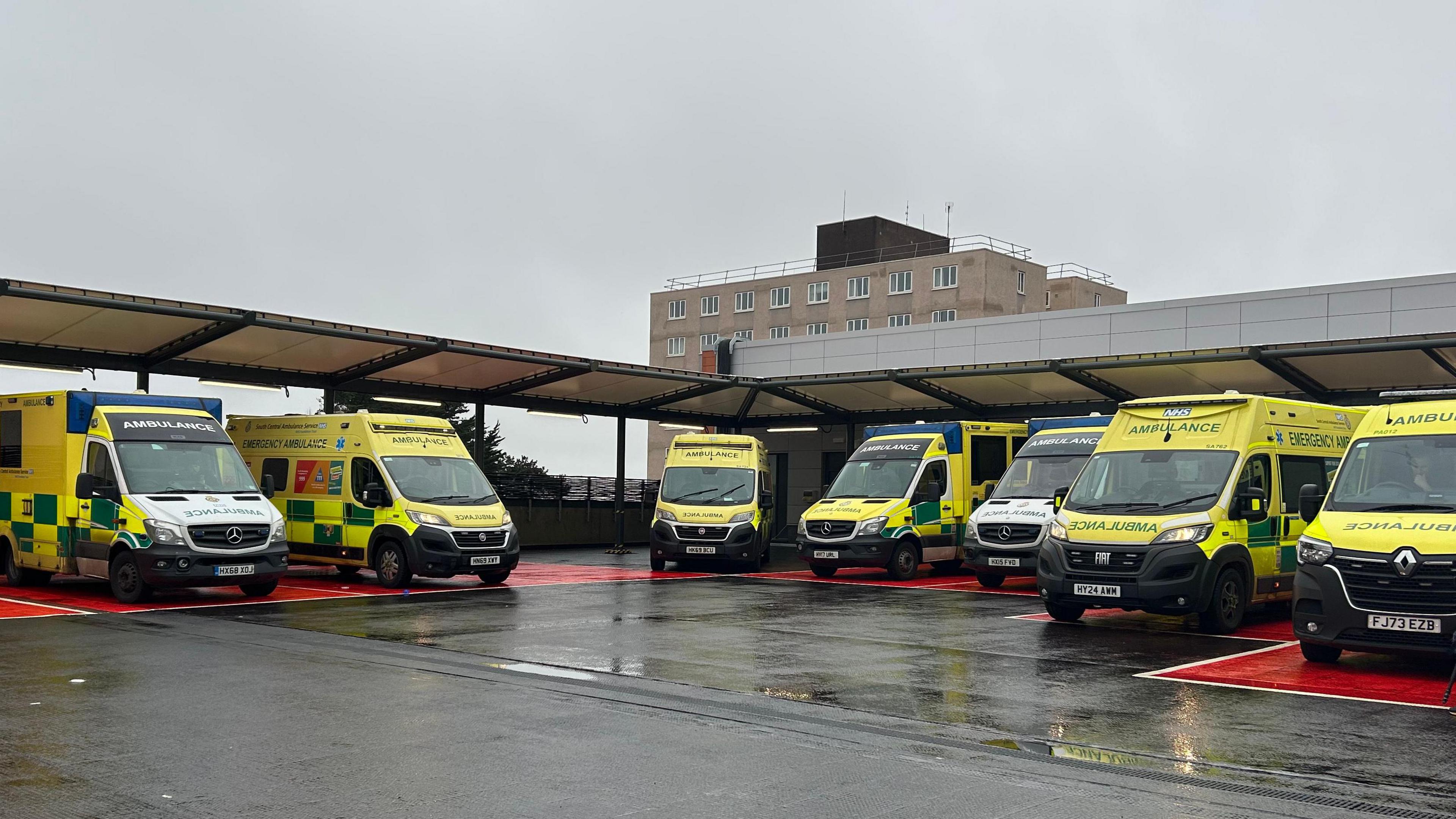 Seven ambulances parked outside QA hospital - they all face outwards so their backs are under the metal canopy.