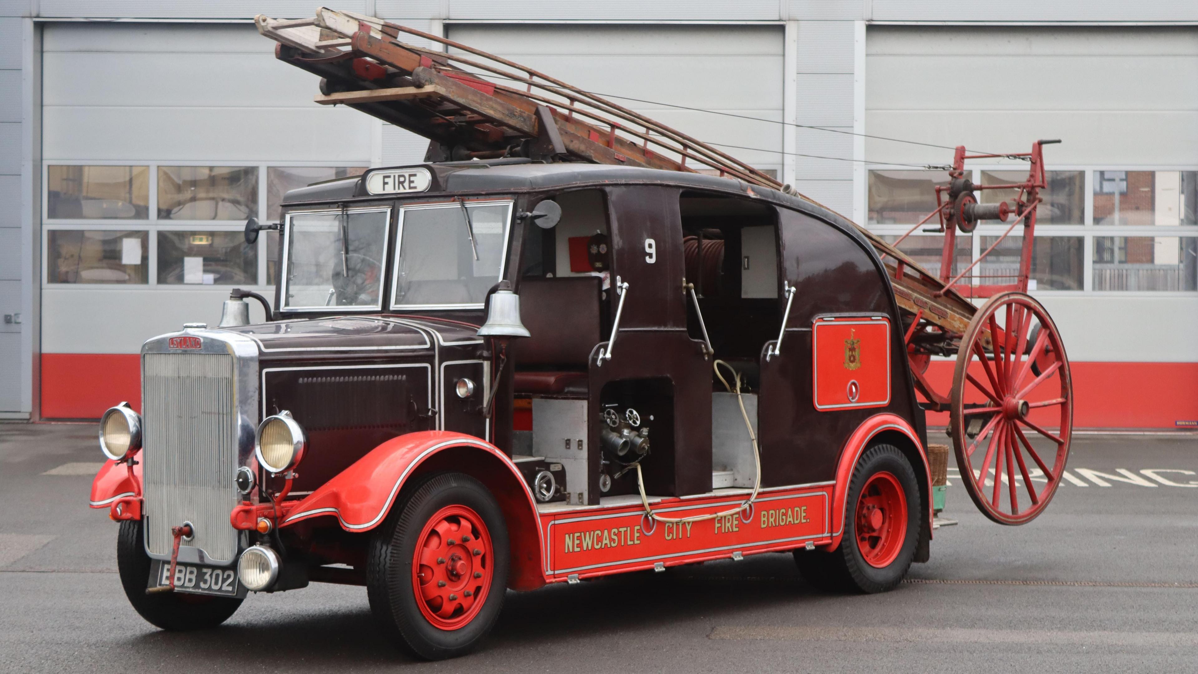 An old fire engine stands in front of a modern fire station. The vehicle has a black body with a flat, two-panelled windscreen and a roof which slopes down at the back. There are silver bells approximately where wing mirrors would usually sit and no doors. The wheel arches, footplate and hub caps are red and along the bottom are the words Newcastle City Fire Brigade. On the roof is a red ladder with a lever and wires attached from the rear. There is a large red wagon wheel at the foot of the ladder.