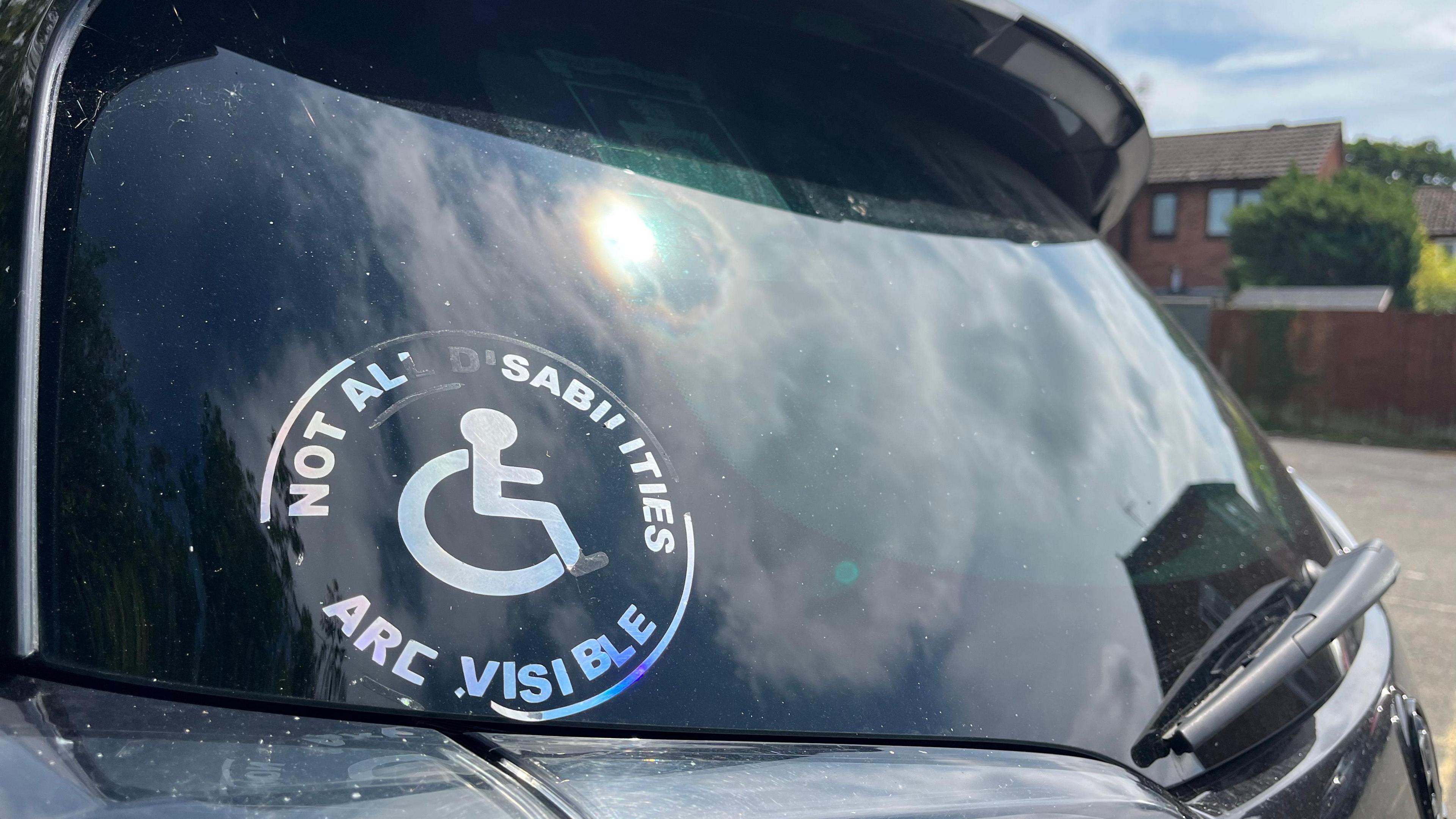 Close up photo of a holographic sticker on the rear window of Mr Tolhurst's car. It has the symbol of a person using a wheelchair in the centre, with the words 'Not all disabilities are visible' around it. 