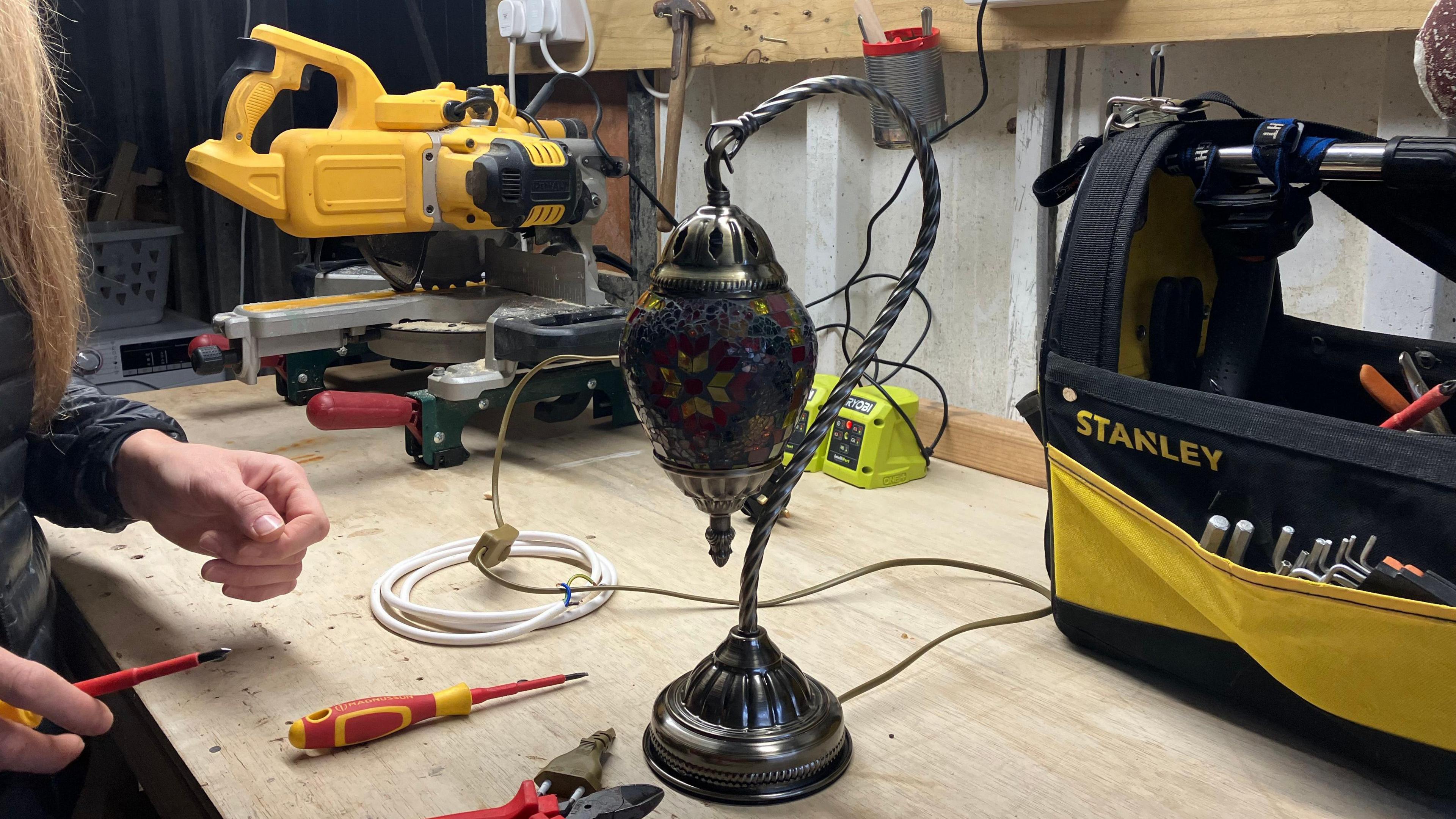 A vintage lamp sits on a work bench, as Sarah holds a screwdriver next to it.