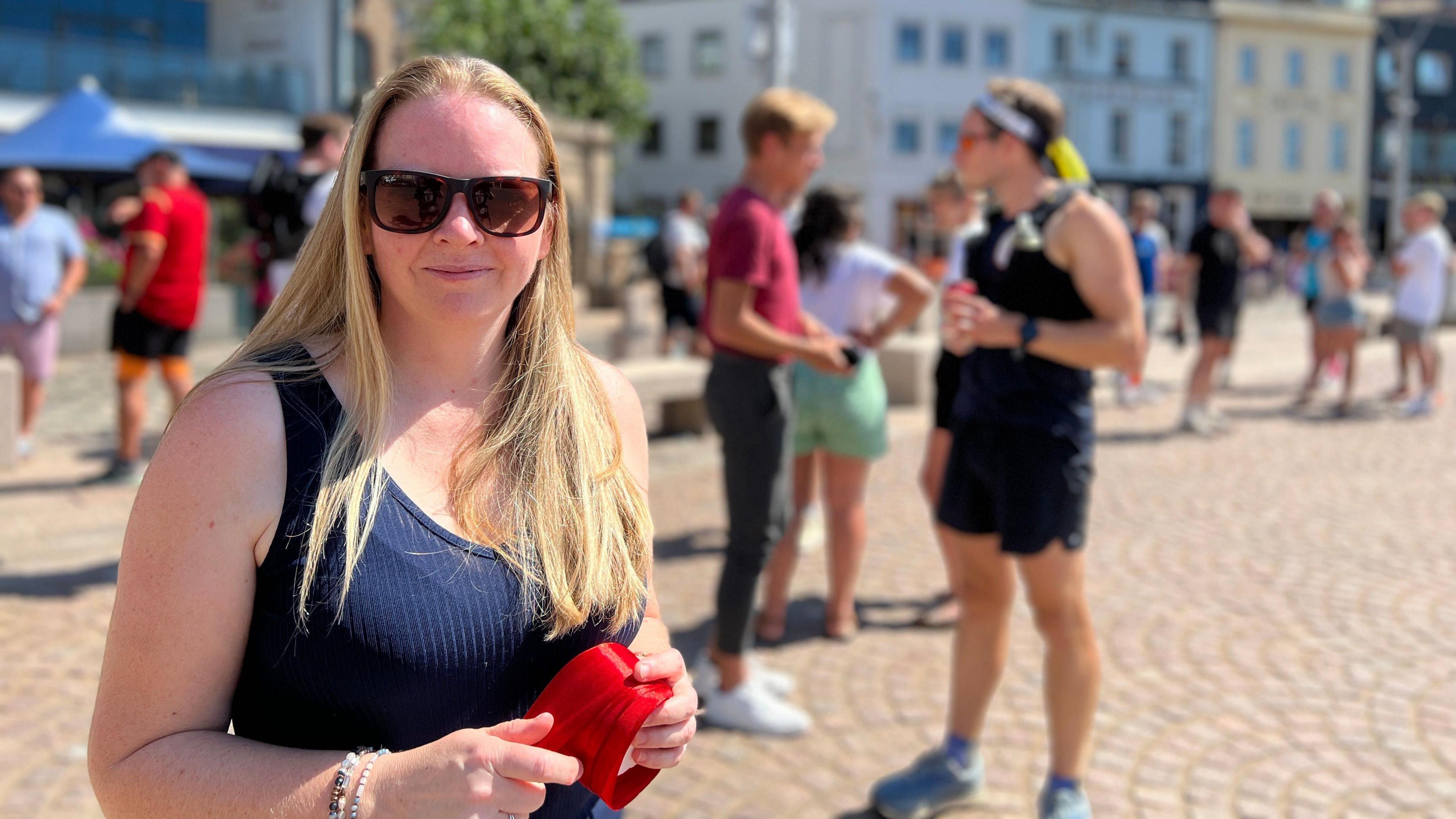 Niamh has long hair stands in a sunny public square holding a red object. Behind her, people walk and talk near buildings under a clear blue sky