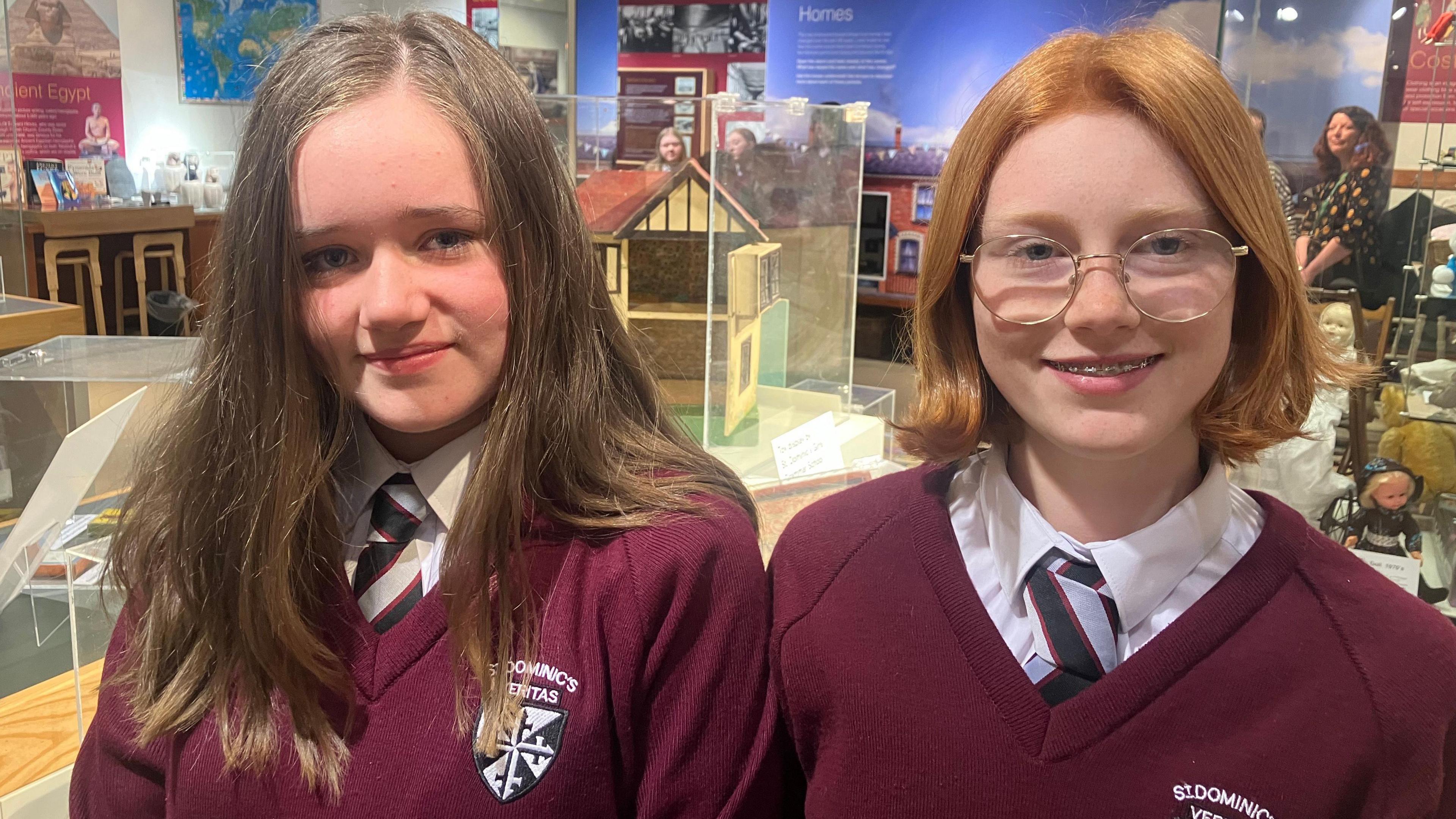 Two girls wearing school uniform of maroon v-neck sweater over a white shirt and striped black, red and silver tie. Aoife on the left has long brown hair and Olivia on the right has an auburn bob and round glasses.