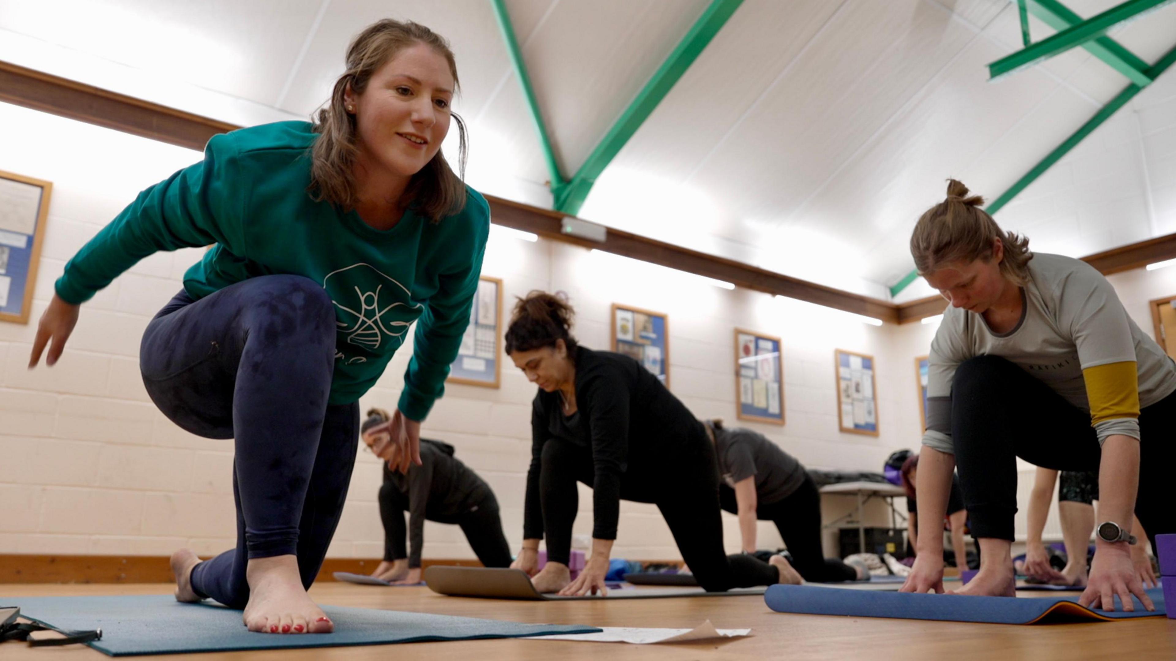 Yoga teacher Hannah Fraser teaches a yoga class. With brown hair, a green jumper and navy leggings Hannah strikes a yoga pose in the foreground while her students follow behind her. 