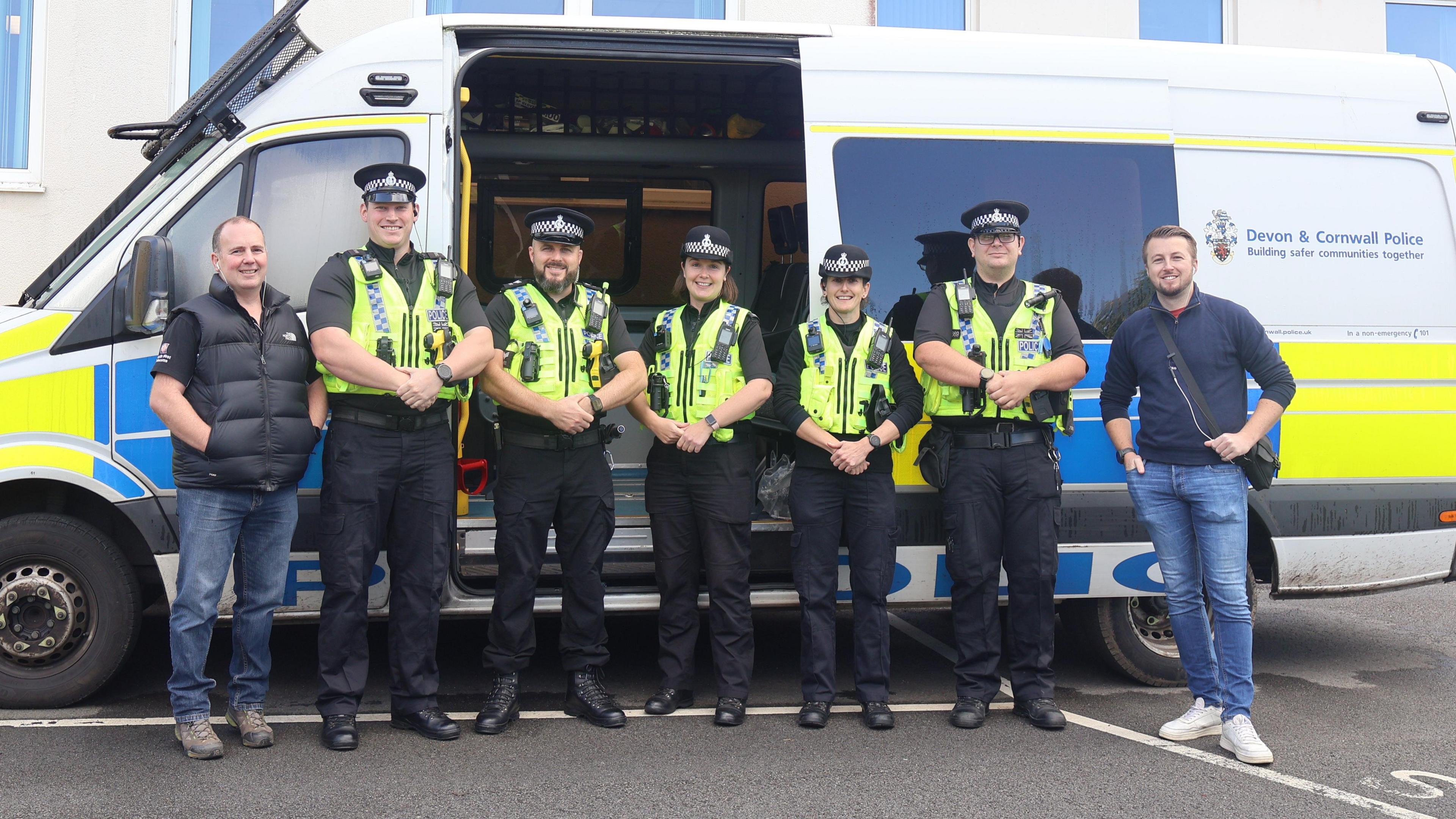 Seven police officers stood in a line in front of a police waggon, which has one of its side door open. The five officers in the centre have high visability uniforms on, which are black with a bright vest. The officers on each end of the row are in plain clothes, wearing casual jeans, T-shirts and jumpers. 