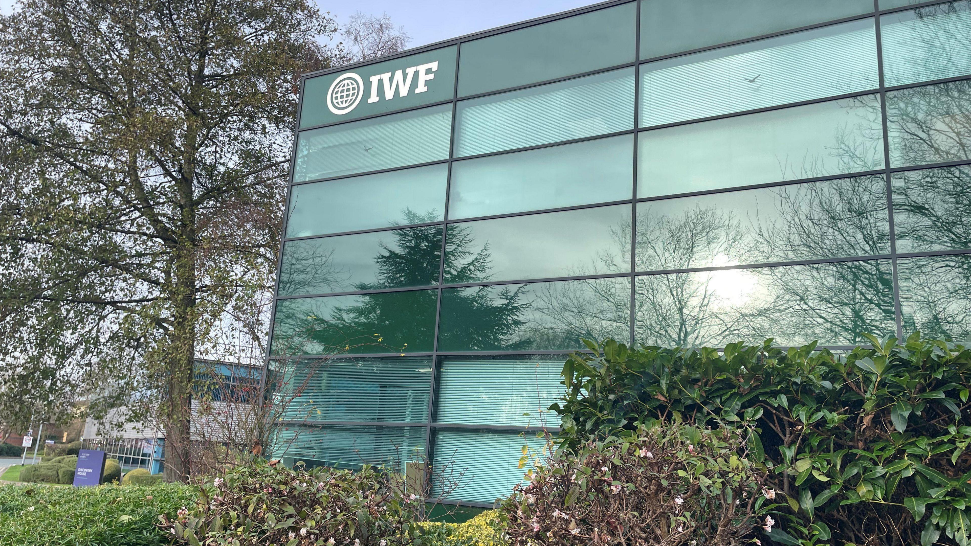 Glass-fronted office block with IWF and a globe sign written in the top-left corner, around the office are other buildings and trees and bushes.