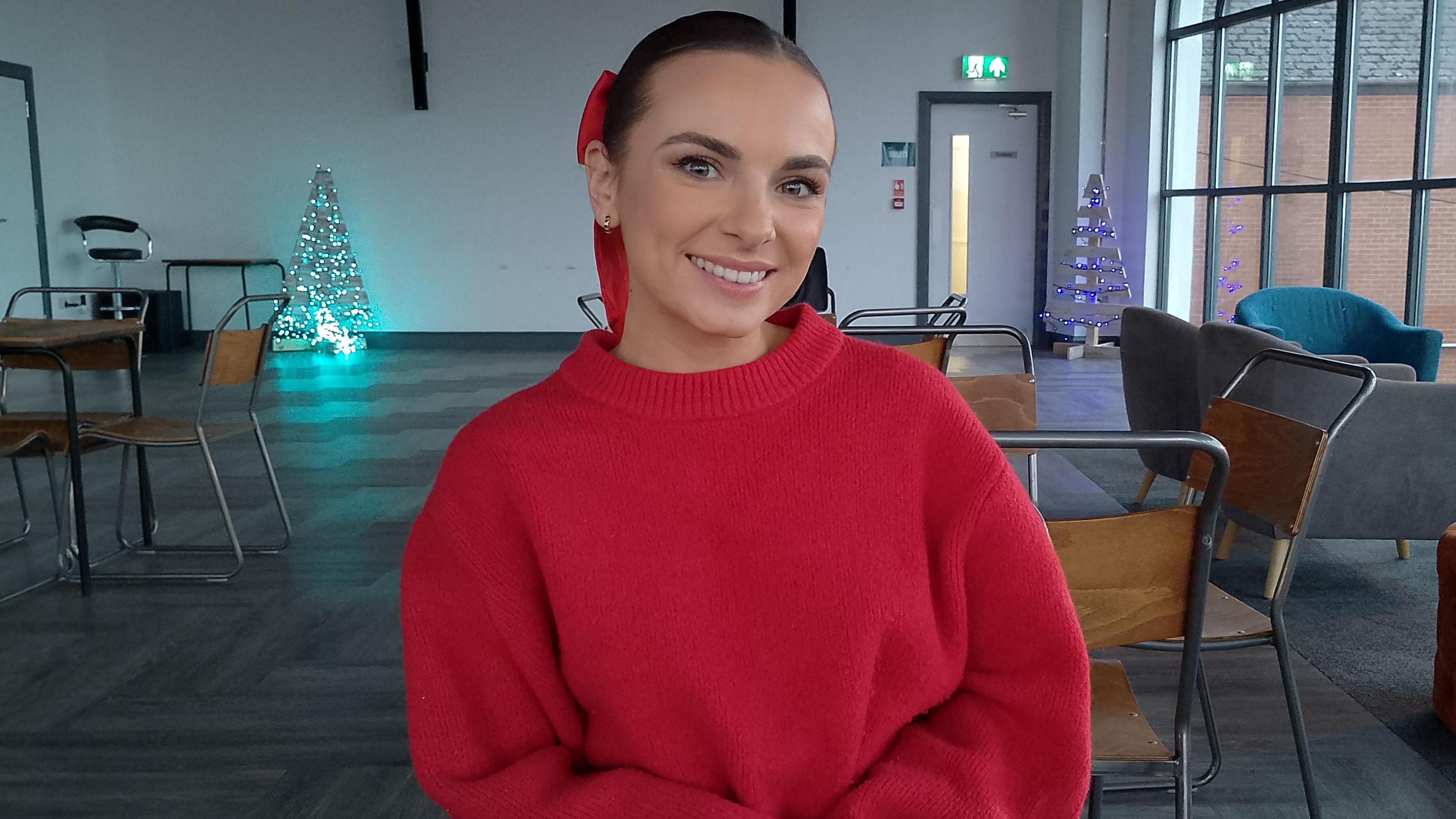 Amber Harrison is wearing a red jumper and has a red bow in her brown hair. She's sitting in a large room, with some Christmas tree decorations in the background. 