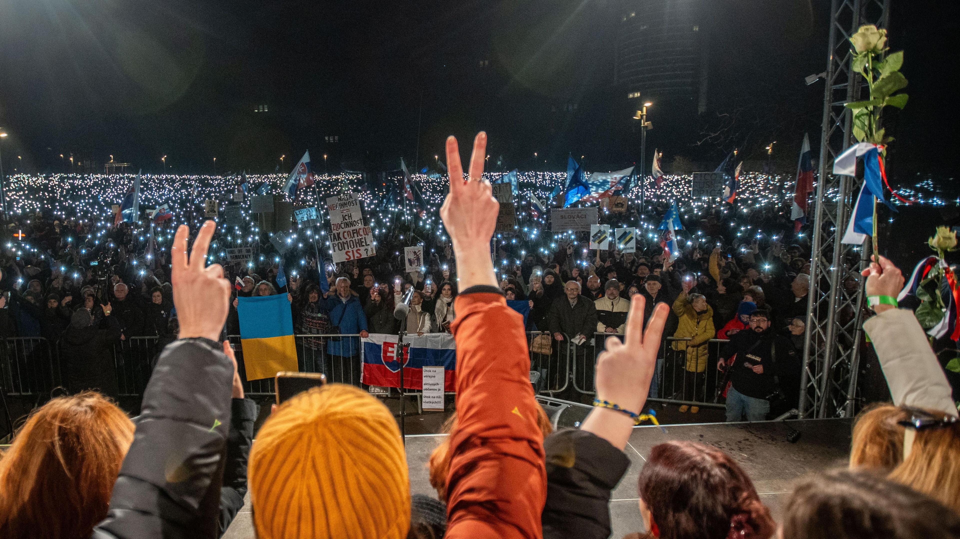 An evening protest in Bratislava, where several demonstrators with their backs to the camera give victory salutes and one holds up a rose. They face a much larger group of protesters behind barriers holding banners and Slovakian and Ukrainian flags