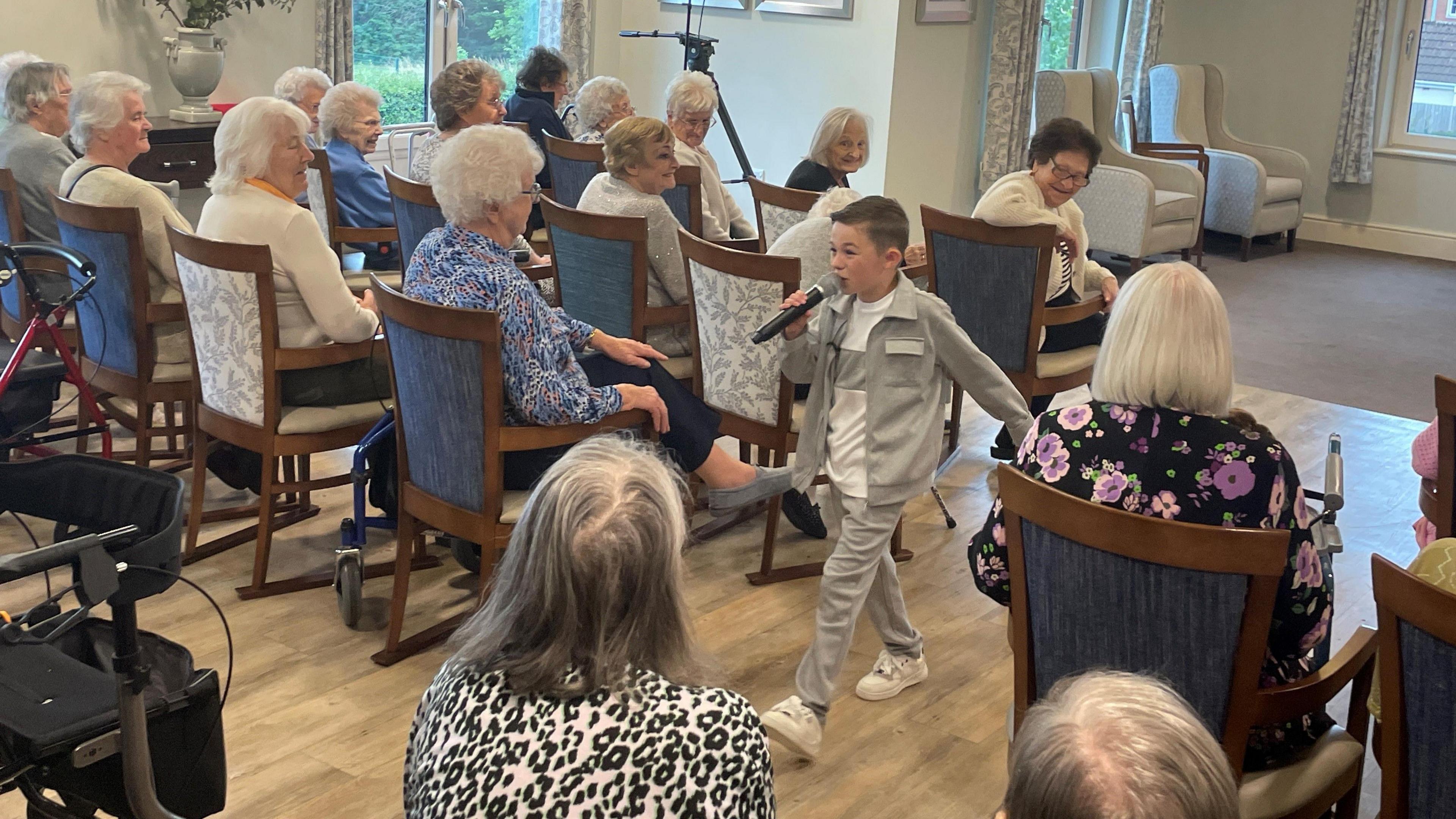 Lucciano wears a grey tracksuit and holds a microphone as he sings and walks out into the audience of seated care home residents.