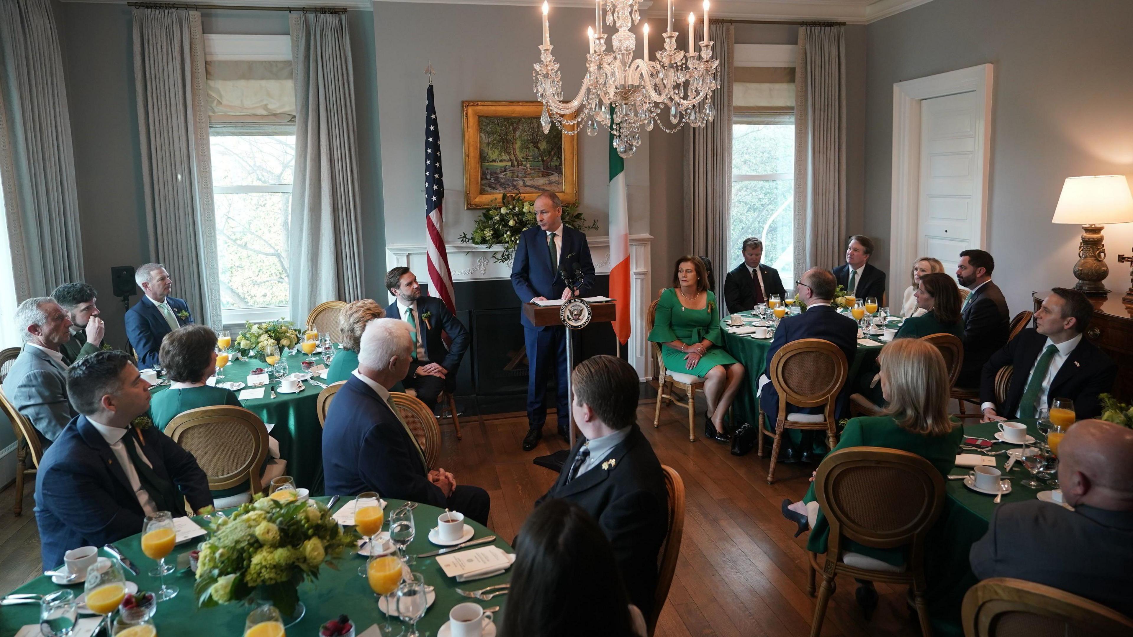 Micheál Martin  stands between a US and Irish flag, in a suit, in a room full of round tables with delegates sat around them 