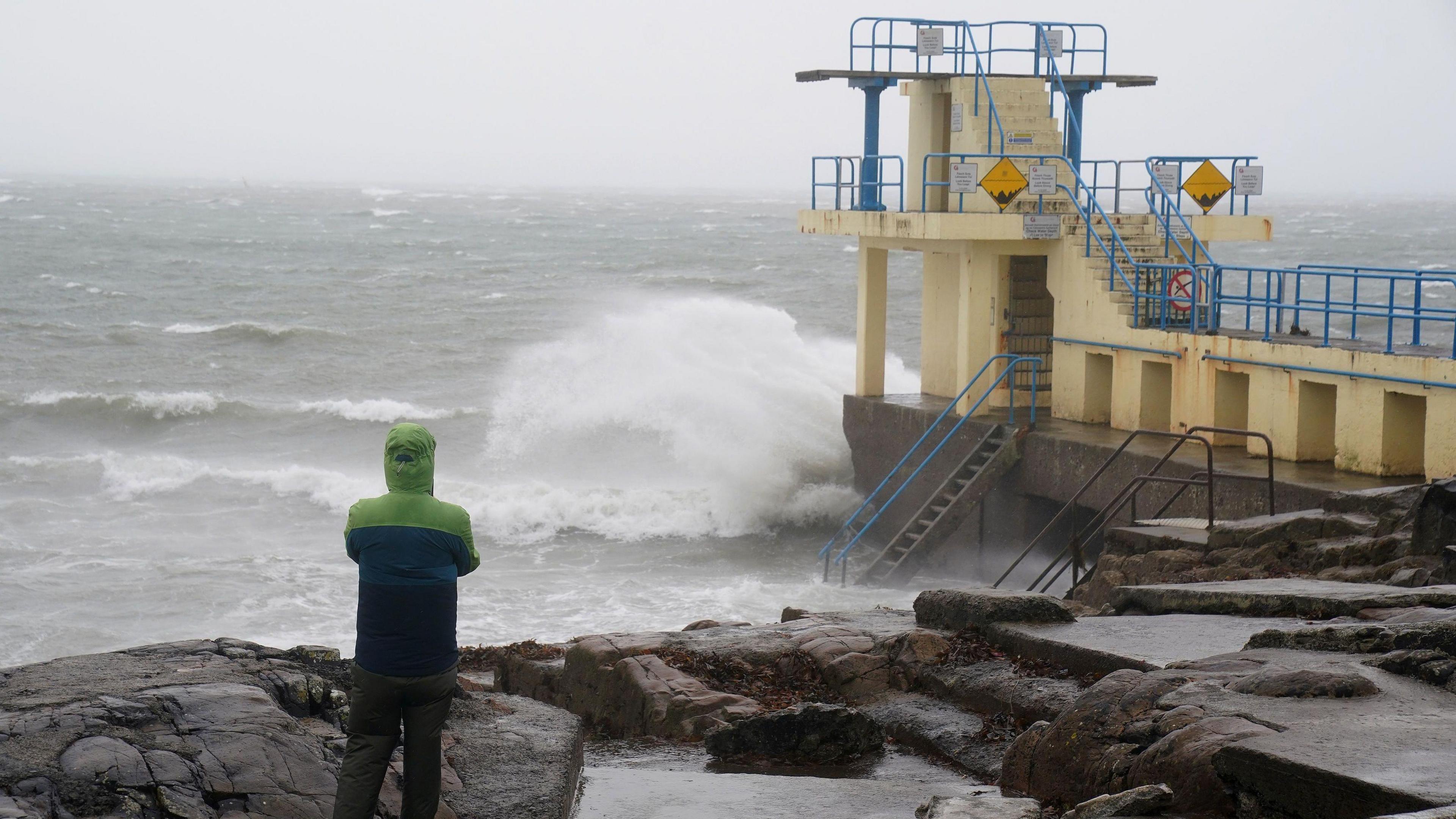 A person wearing a green and blue coat stands with their back to the camera watching waves crash over a bathing spot on the coast.