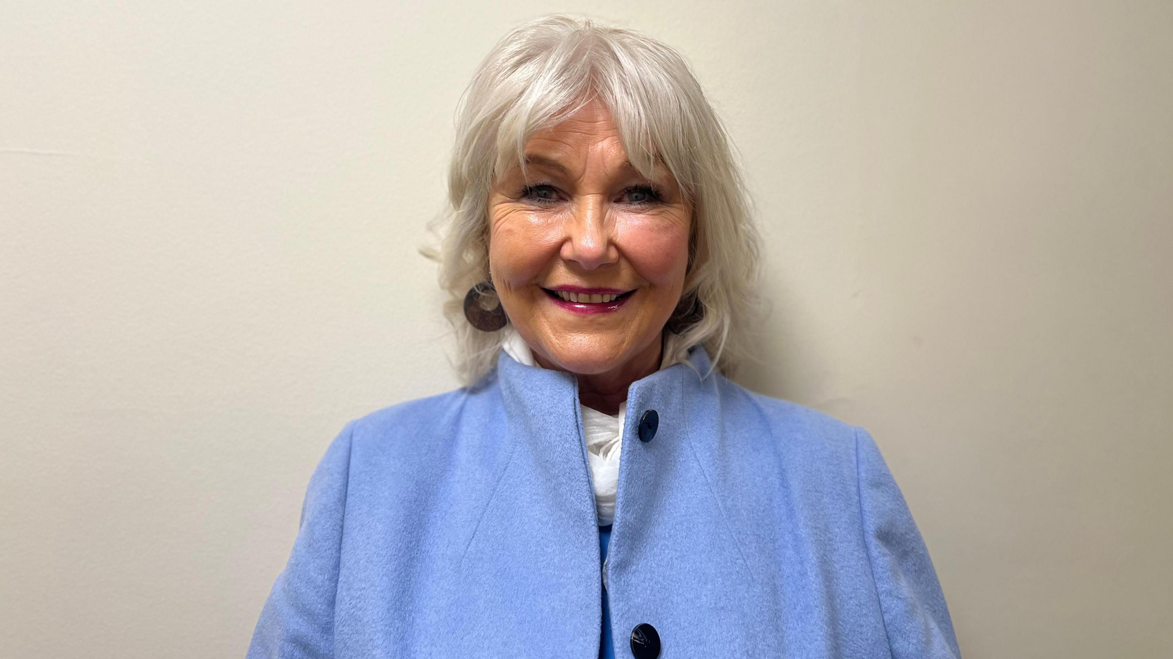 Victims campaigner Margaret McGuckin is standing in front of a white background. She is wearing a blue coat with black buttons. She has grey hair and is wearing a white scarf. 