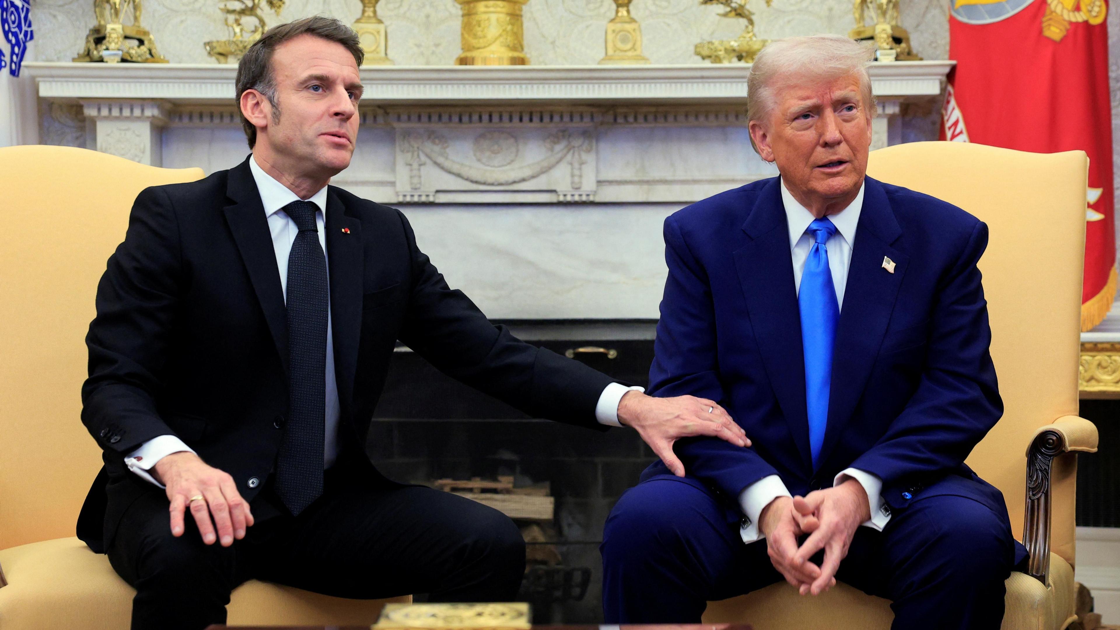 Macron placing his hand on Trump's arm as they take questions from reporters. Both men are wearing dark suits with matching ties.