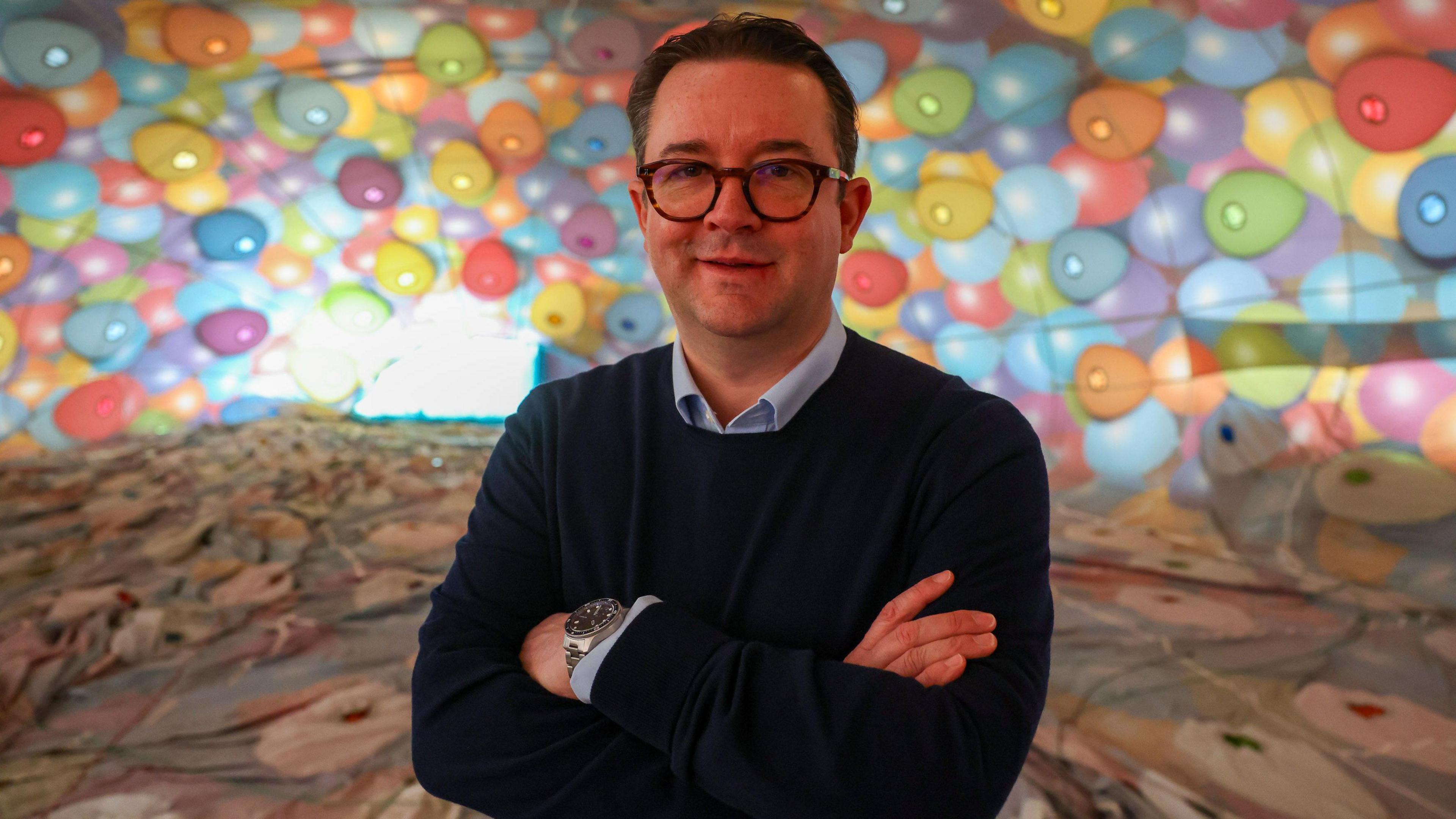 Ben Hardy. He has short dark hair and is wearing glasses with thick black frames. He is standing in front of a hot air balloon envelope which is multi coloured. Ben is wearing a blue collared shirt and a navy blue jumper. He is standing with his arms crossed, looking at the camera and smiling.