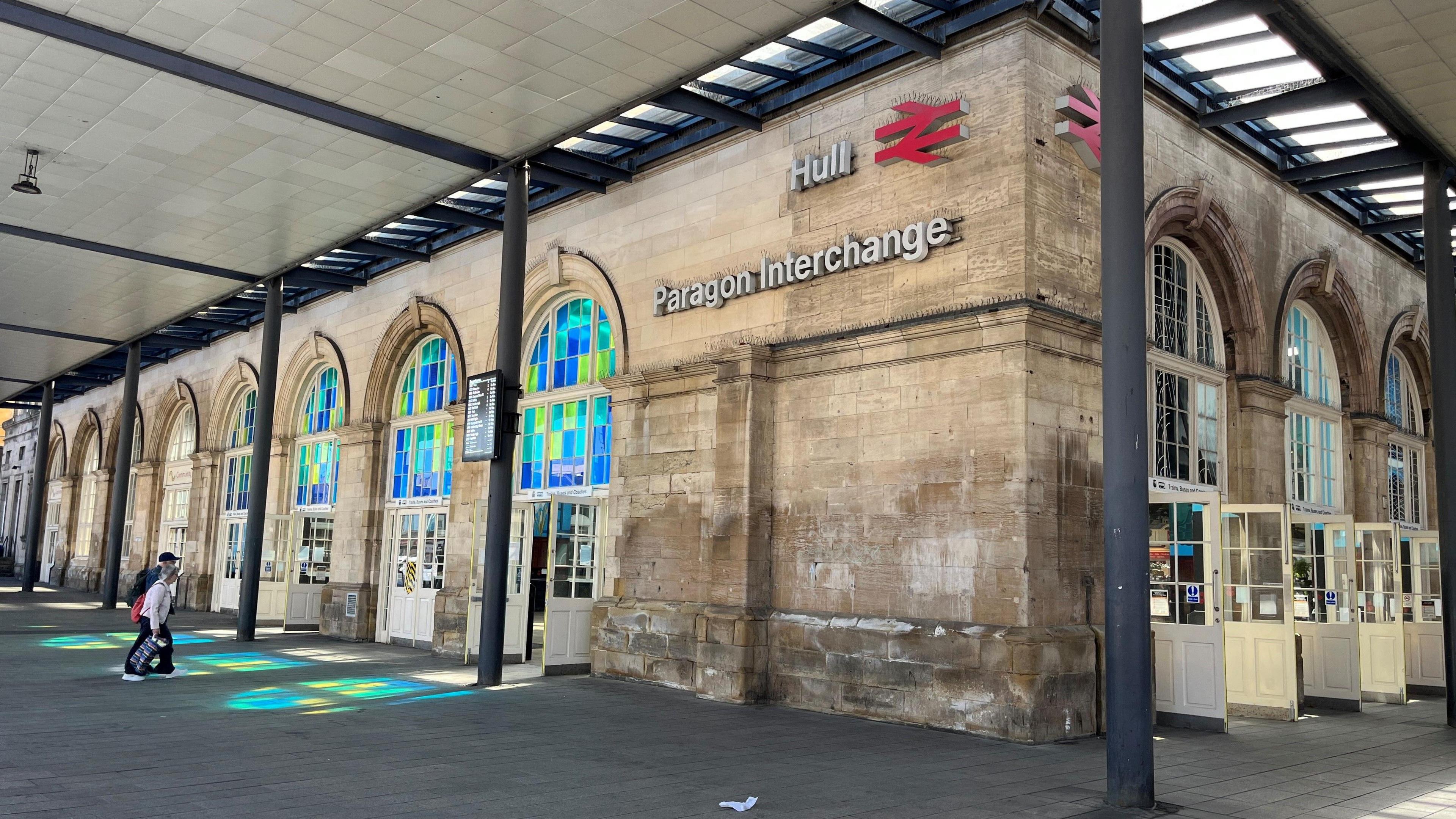 An exterior view of Hull Paragon Interchange. The building has large arched windows with colourful stained glass, white entrance doors and black metal support columns. Above the entrance a sign reads "Hull" with the British Rail logo and "Paragon Interchange". A person is walking past the building, pulling a suitcase.