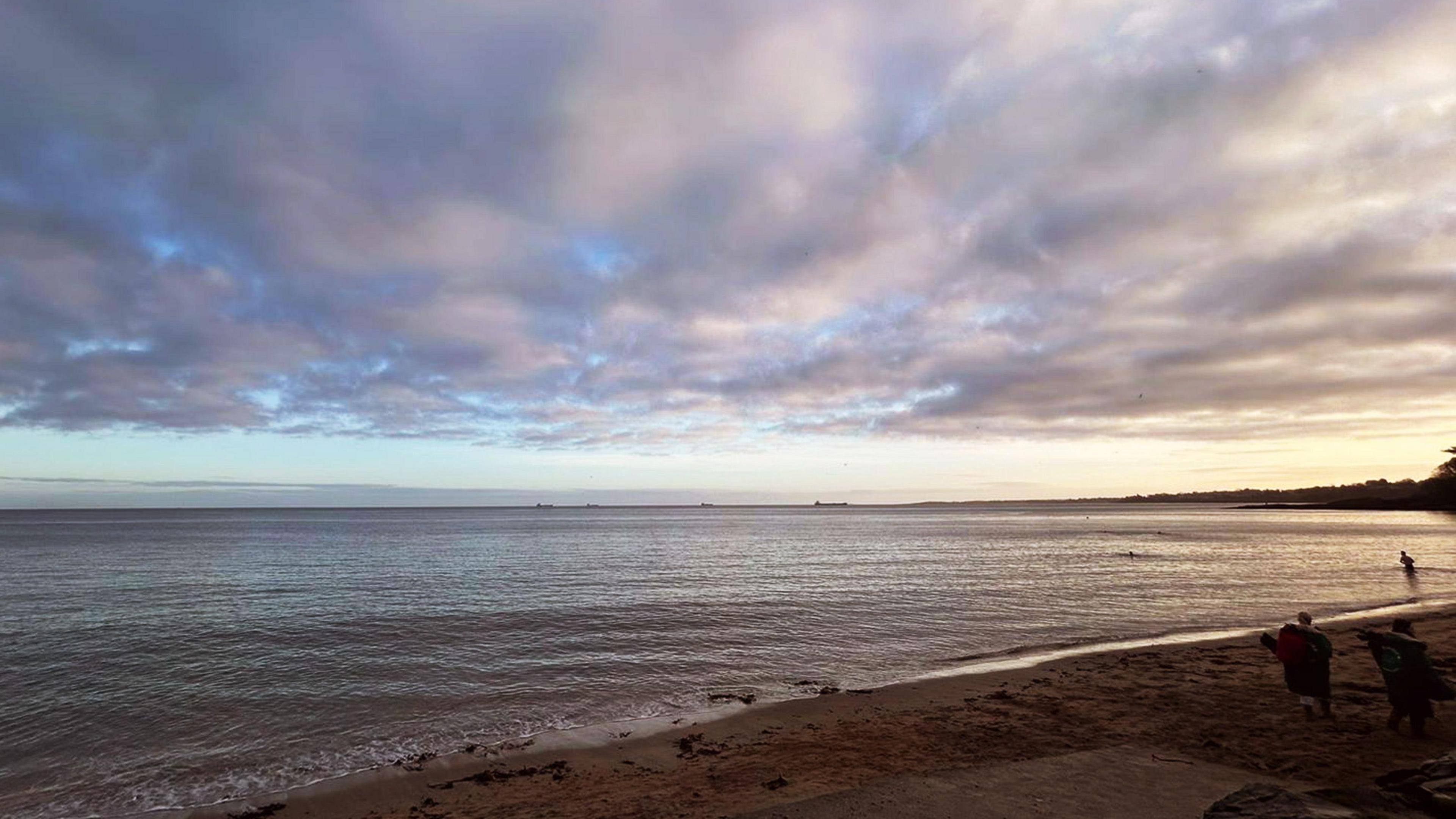 A sky full of broken cloud over the sea at Helen's Bay beach where a small section of sand can be seen in the lower right hand corner. Two people in the distance are carrying deck chairs across the sand, while a man further away can be seen entering the water. There is a soft orange light indicating a sunrise. Four ships sit far in the horizon. 