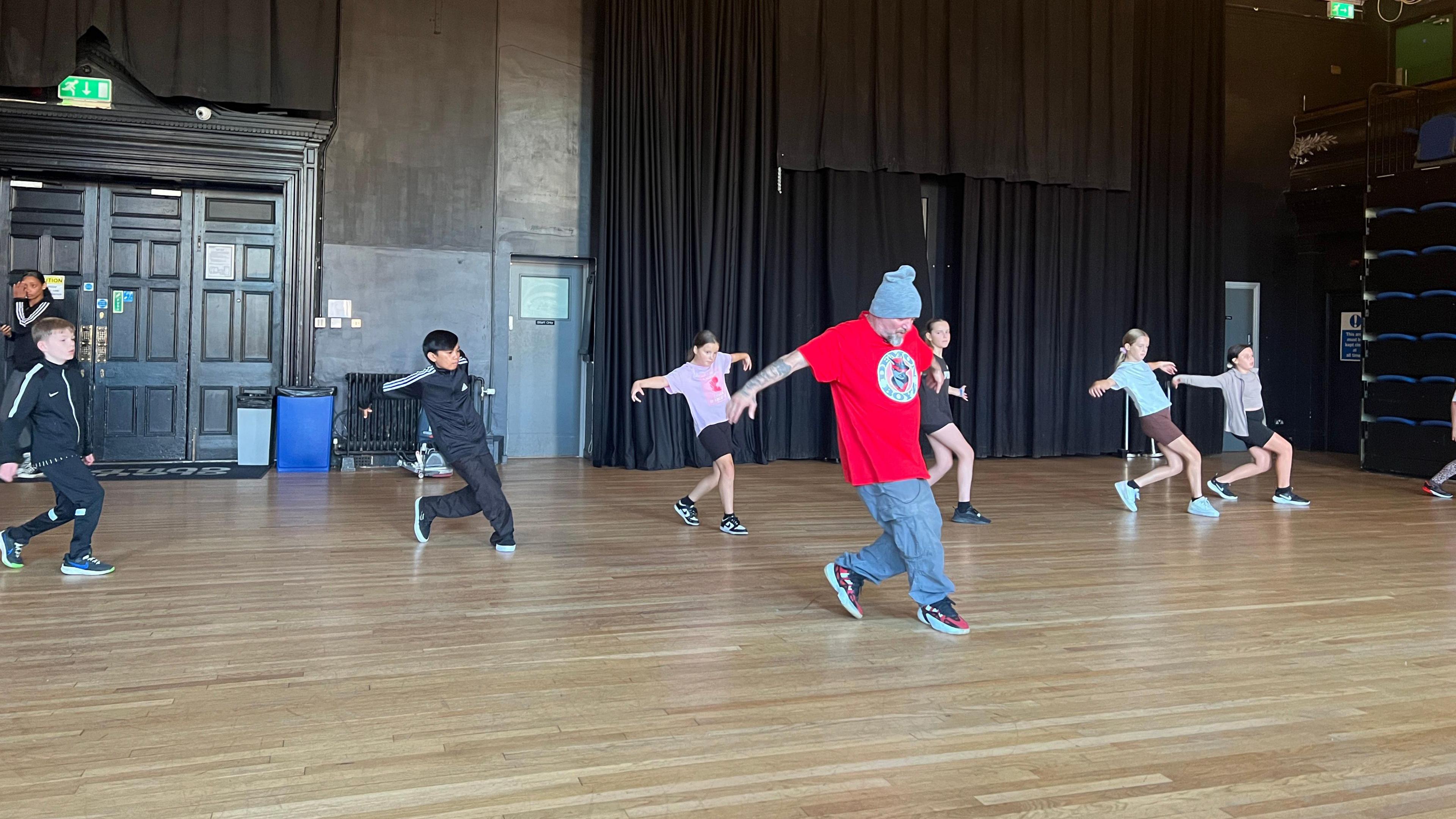 Six children are seen performing break dance moves behind a teacher who is wearing a bright red t-shirt and a beanie hat.