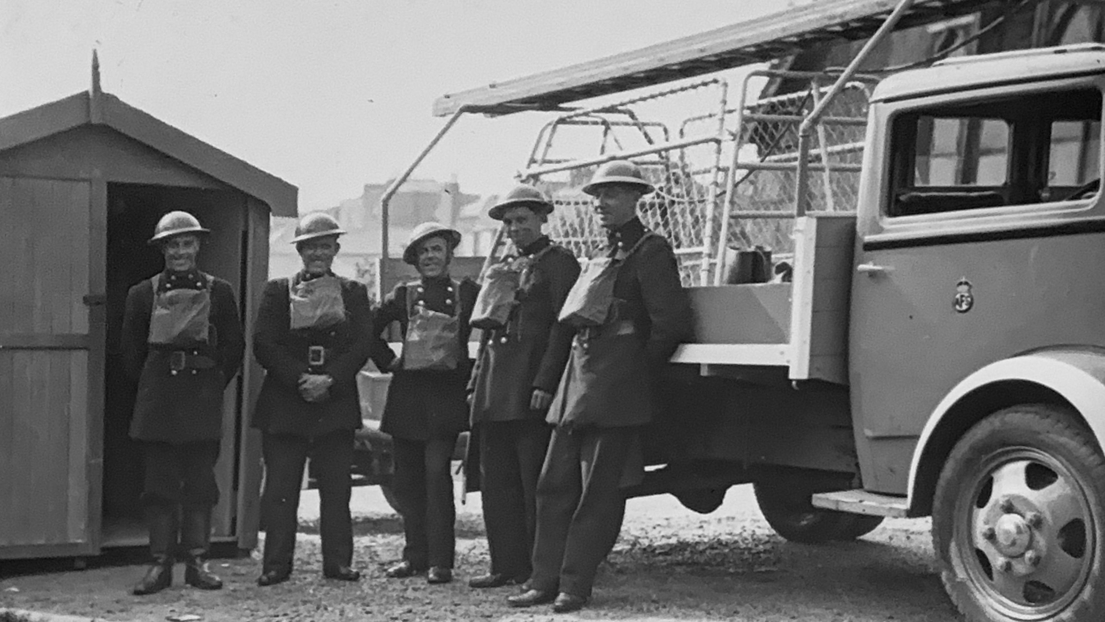 A black and white photo showing firefighters standing alongside a truck, they have tin hats on and bags around their necks. They are all wearing button up jackets and trousers and smiling at the camera.