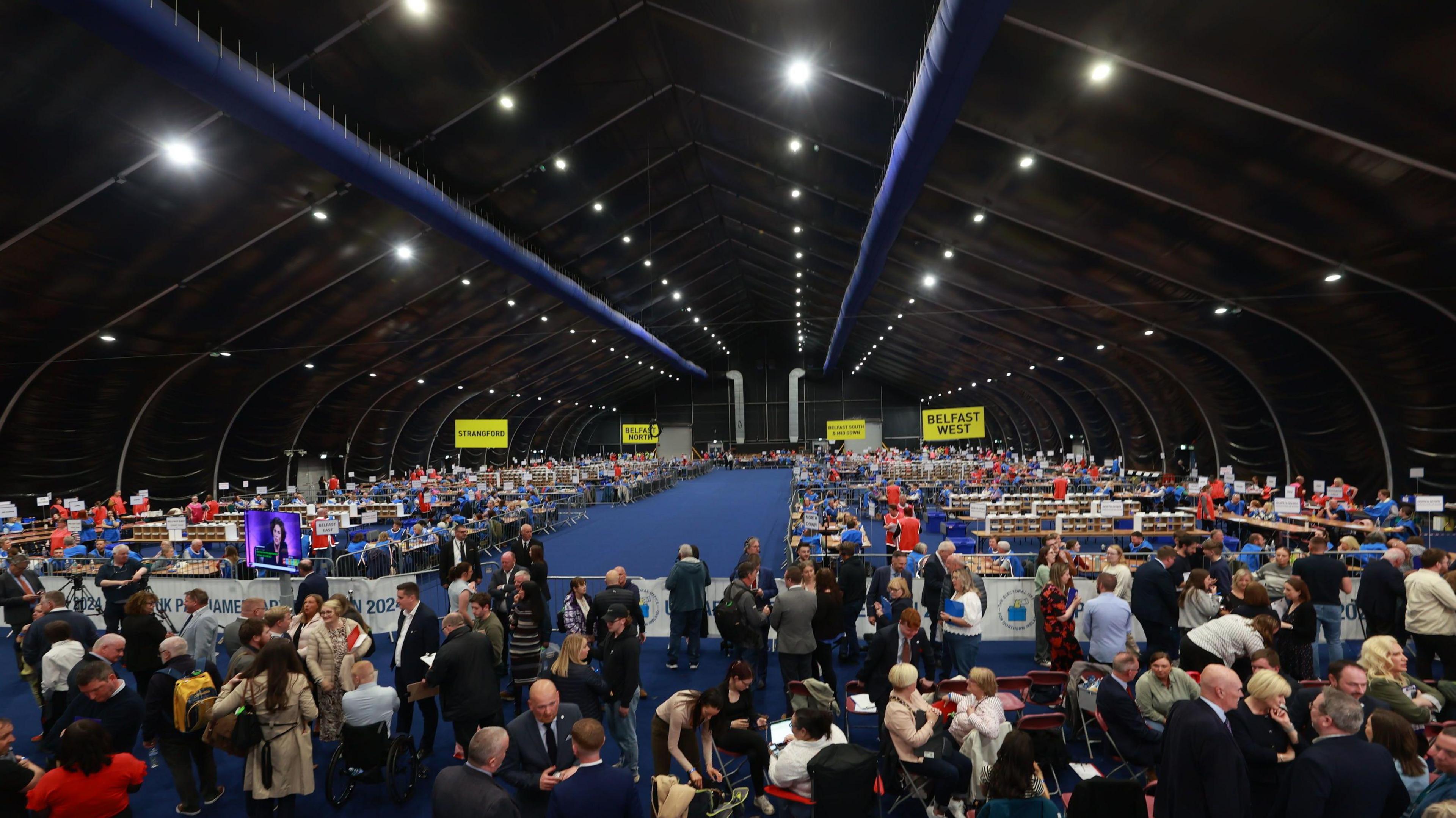 Crowds of people stand on a blue carpet inside the Titanic Exhibition Centre where votes are counted for local elections. There are yellow signs for different constituencies around Belfast. 