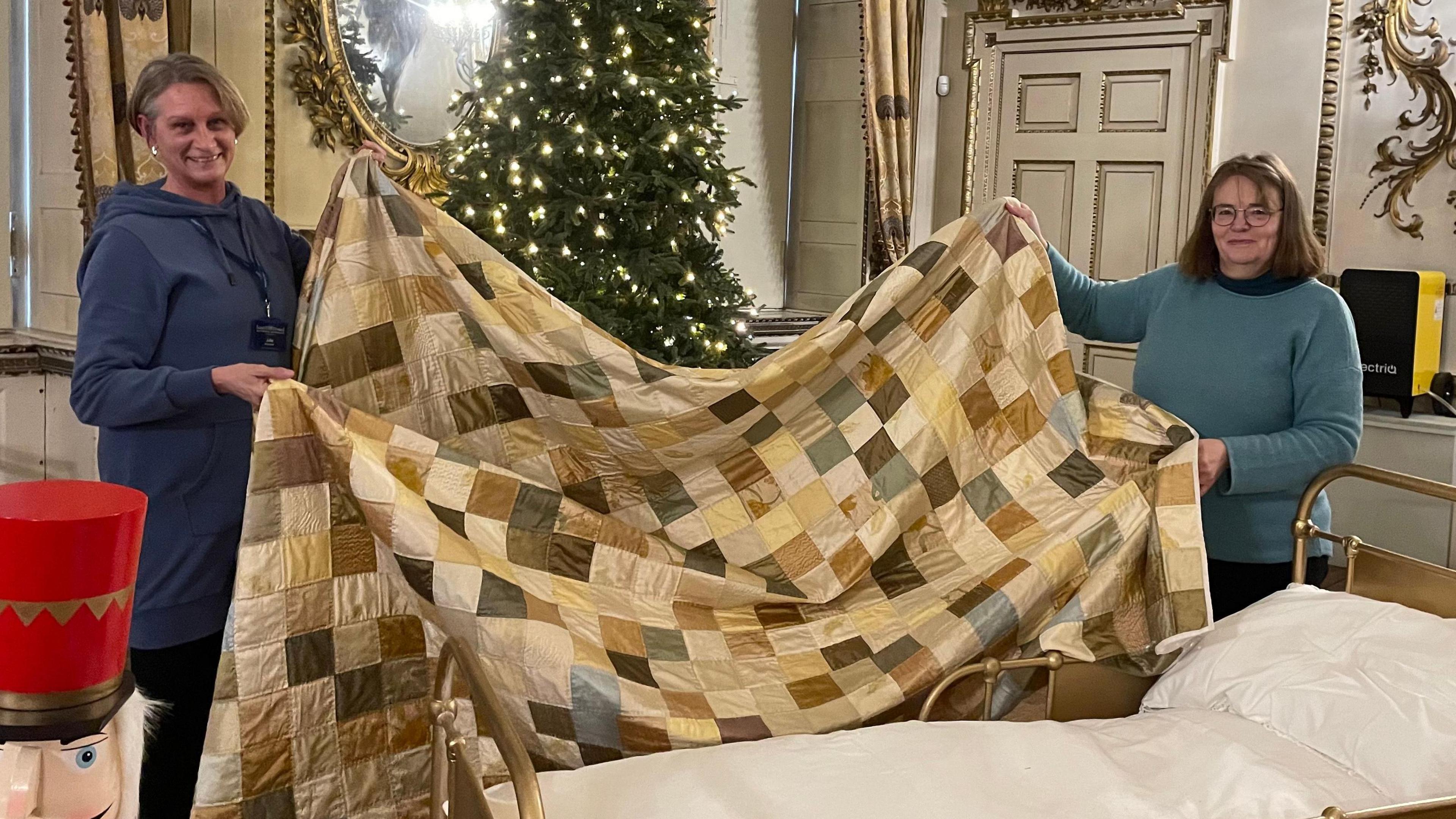 Victoria and Julie are holding a very big quilt, which has been sewn in a check pattern of various different fabrics. They're trying to put it over a small children's bed.