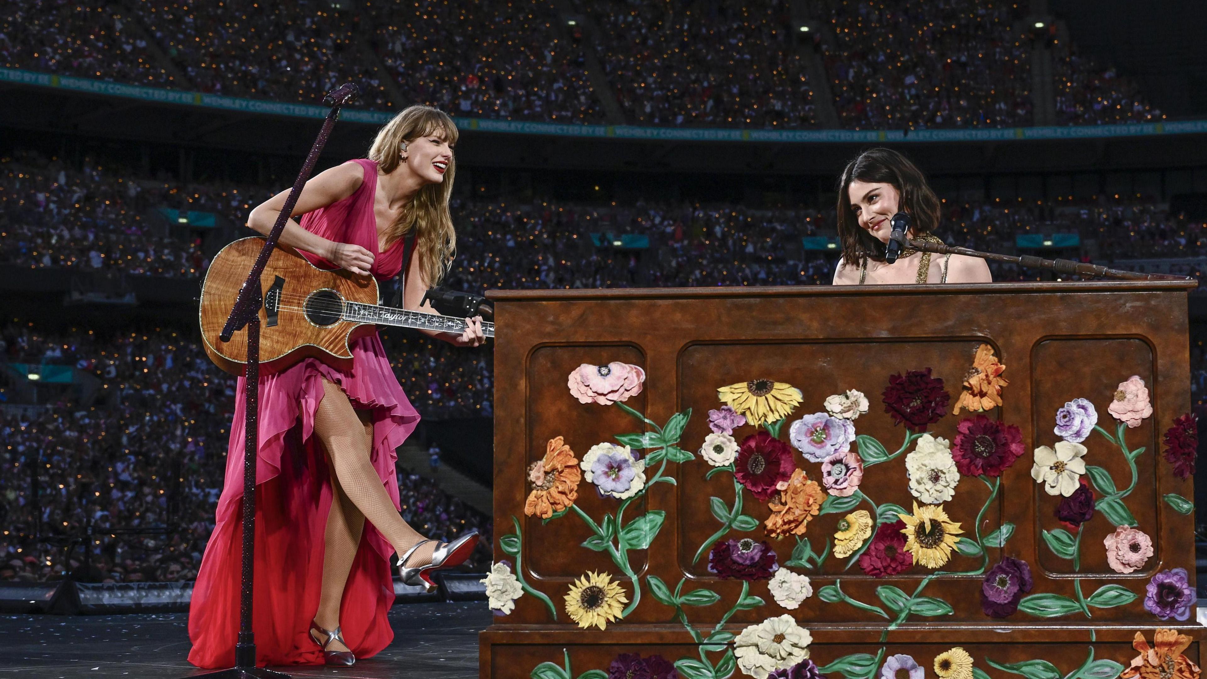 Taylor Swift performing on stage with Gracie Abrams, who is sat at a piano