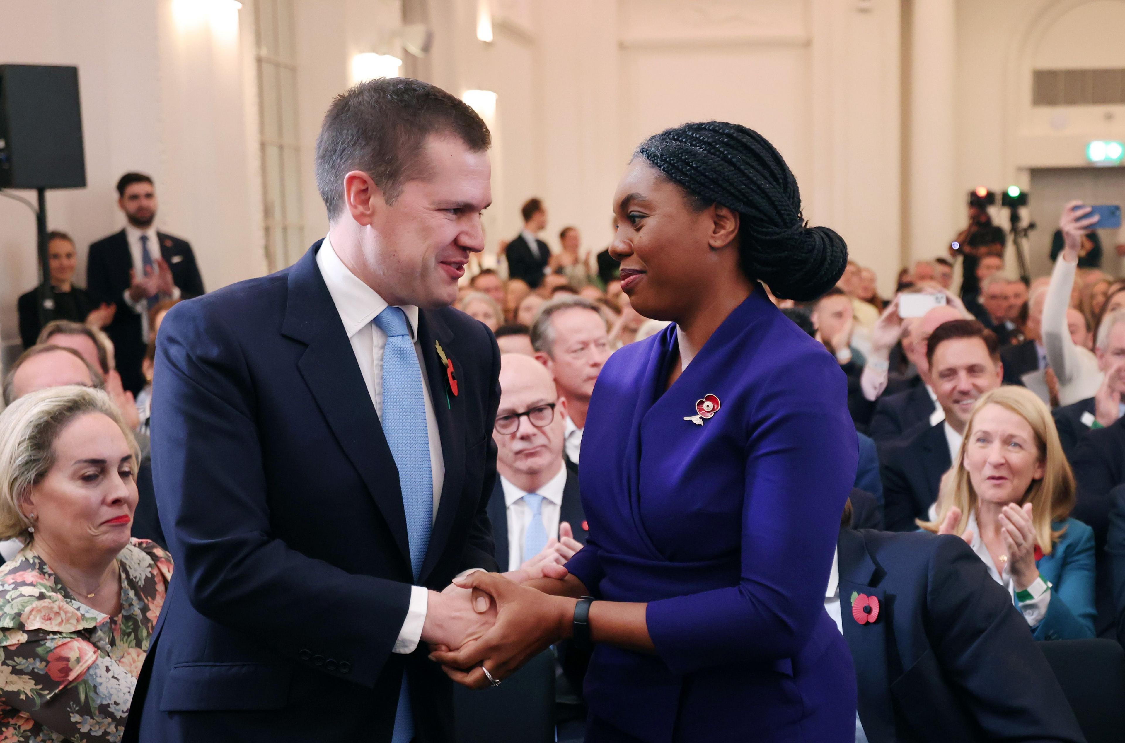 Robert Jenrock, wearing a dark blue suit and white shirt, shakes the hand of Kemi Badeoch, who wears a blue dress suit and has her braided hair back in a bun.