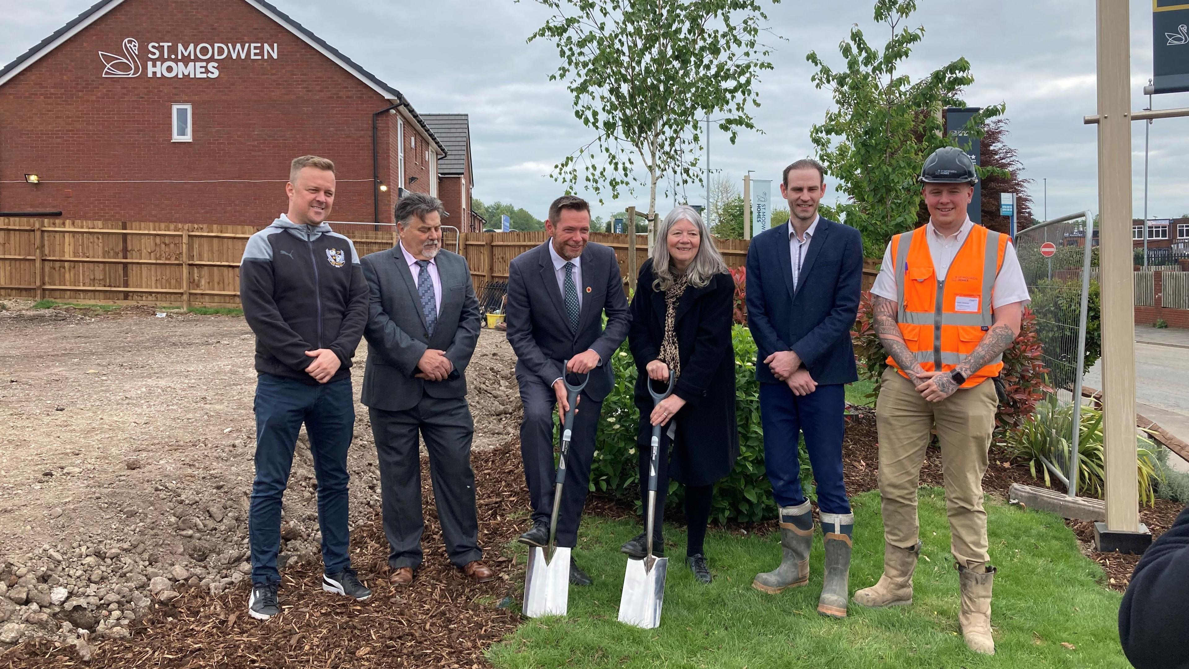 Five men and a woman stand in a line, with one of the men and the woman (both in the centre) holding spades. The man on the right is wearing a hard hat and orange hi-vis.