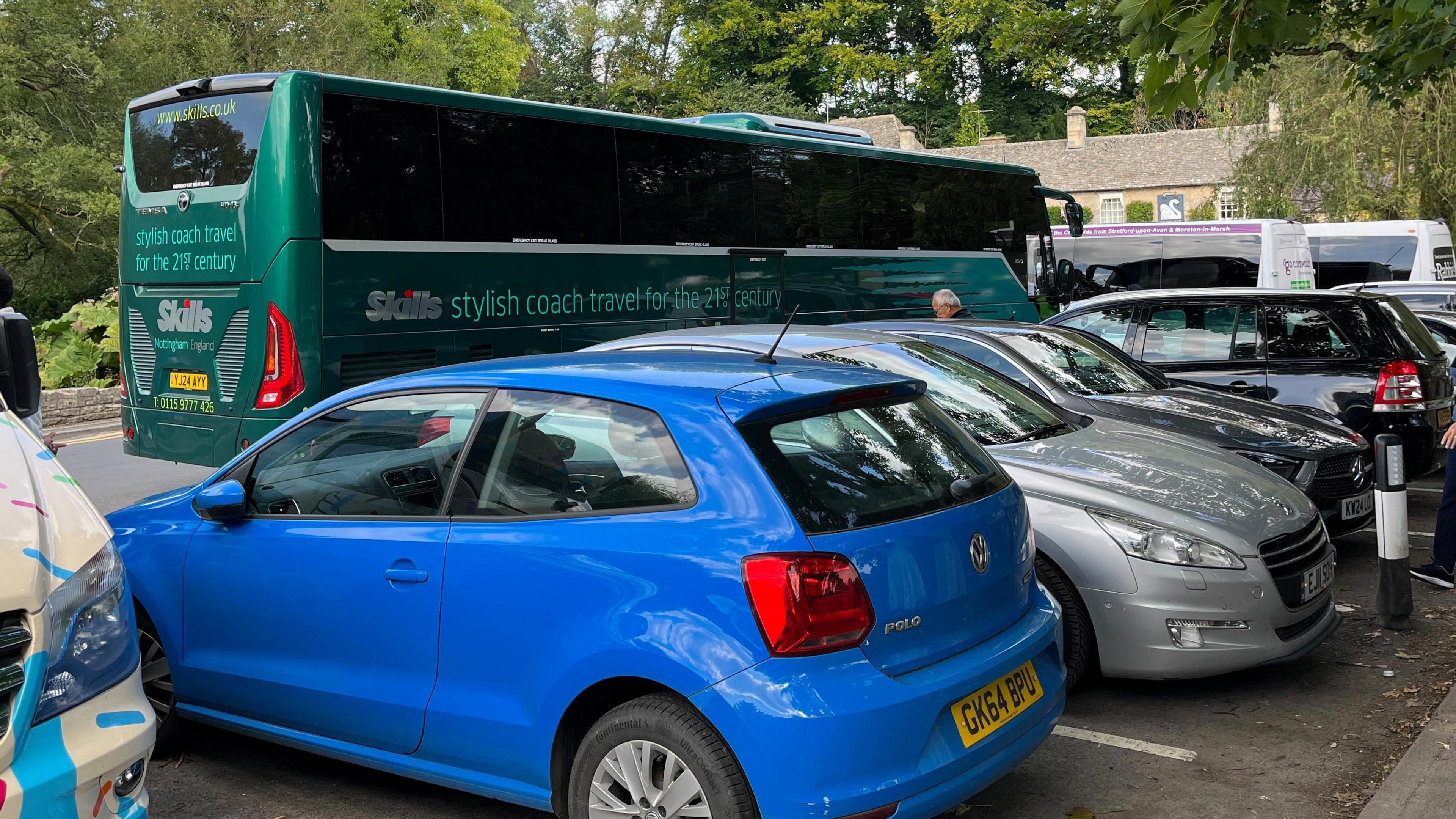 An alternate view of the small parking bay area in Bibury from the car side. There is a row of cars being blocked into bays by the large green coach.