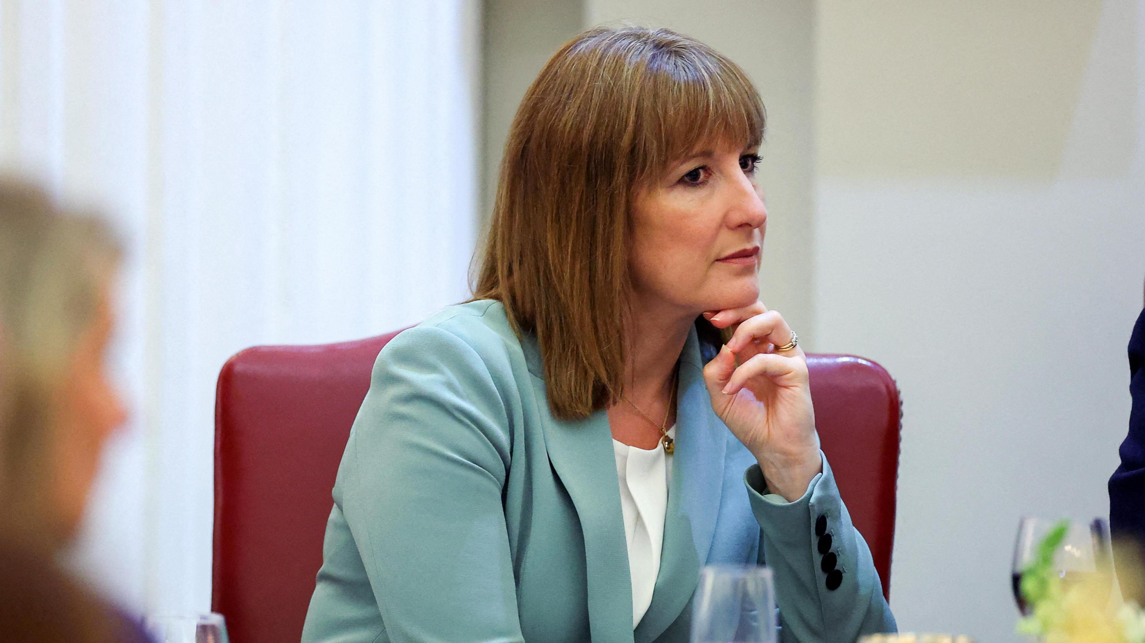 British Chancellor of the Exchequer Rachel Reeves is sat in a red leather chair, in a pale blue blazer. Her chin is resting on her hand.