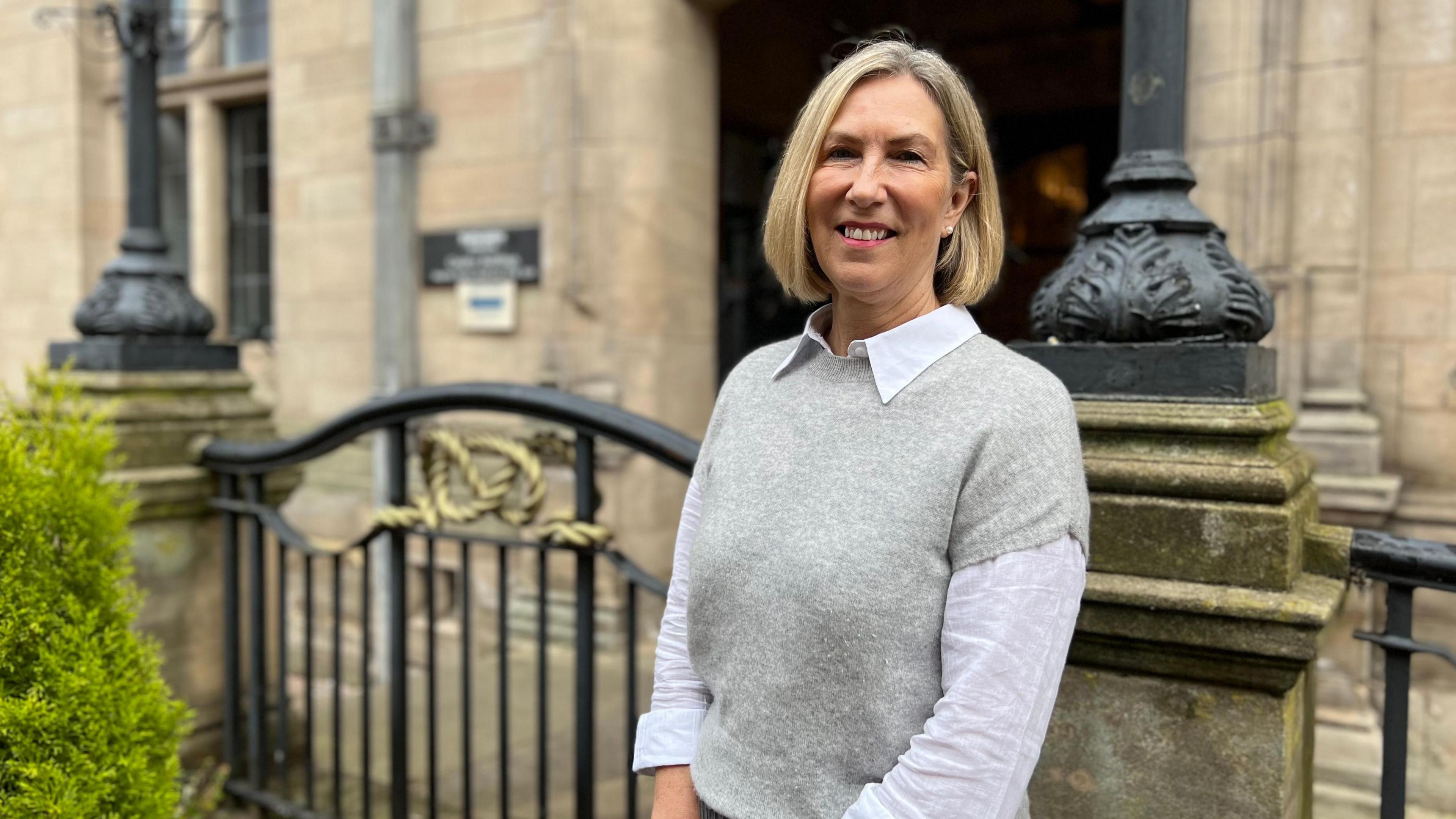 Elizabeth Barnes, a woman wearing a white shirt and grey jumper. She is stood outdoors, in front of a stone building with an ornate black and gold gate.