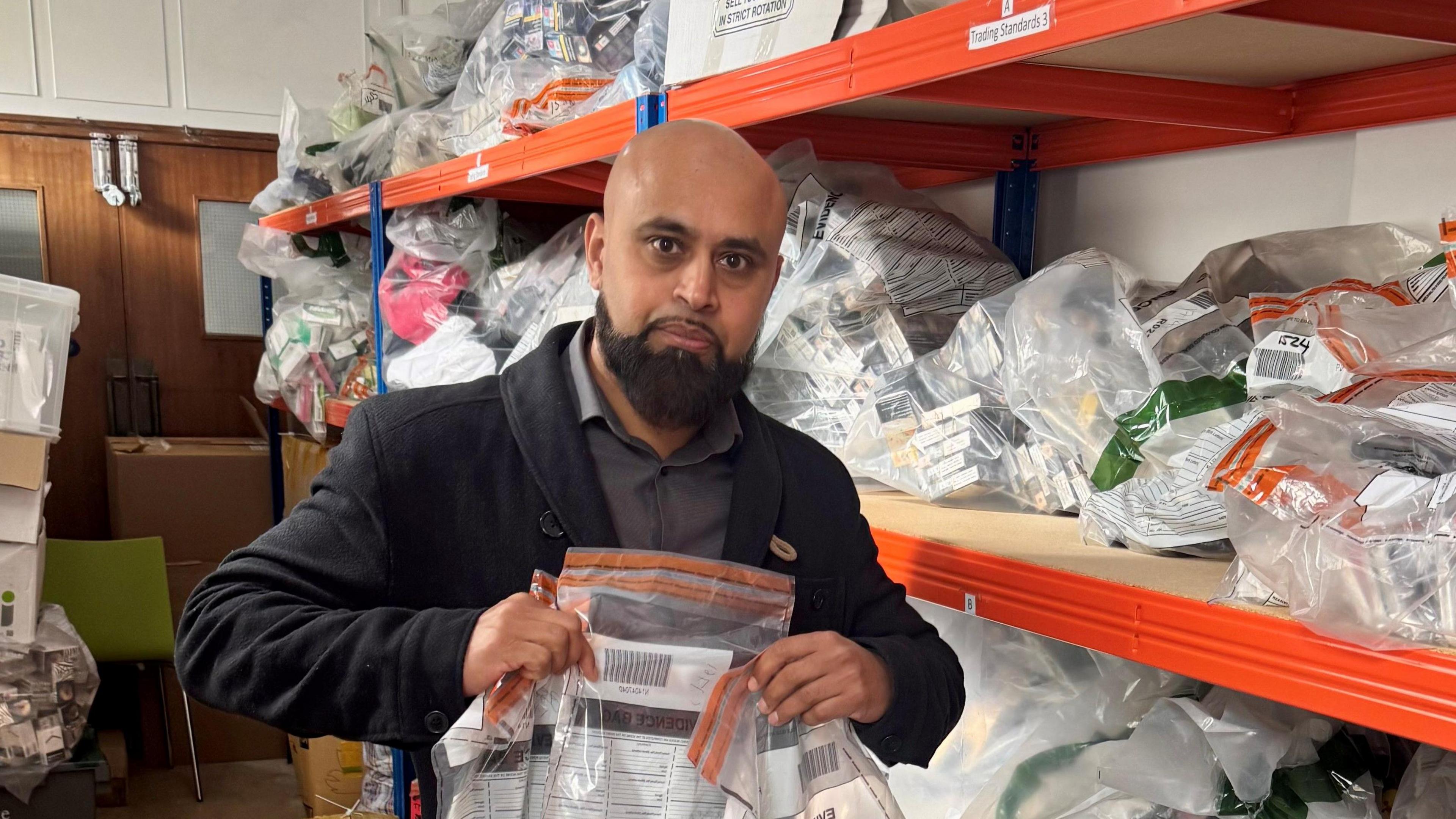 Nazir Ali holds a clear plastic evidence bag up for the camera. He is standing in a room with shelves behind him that are filled with similar evidence bags. Nazir is a bald man, with a thick black beard. He is wearing a black jacket and a grey shirt.