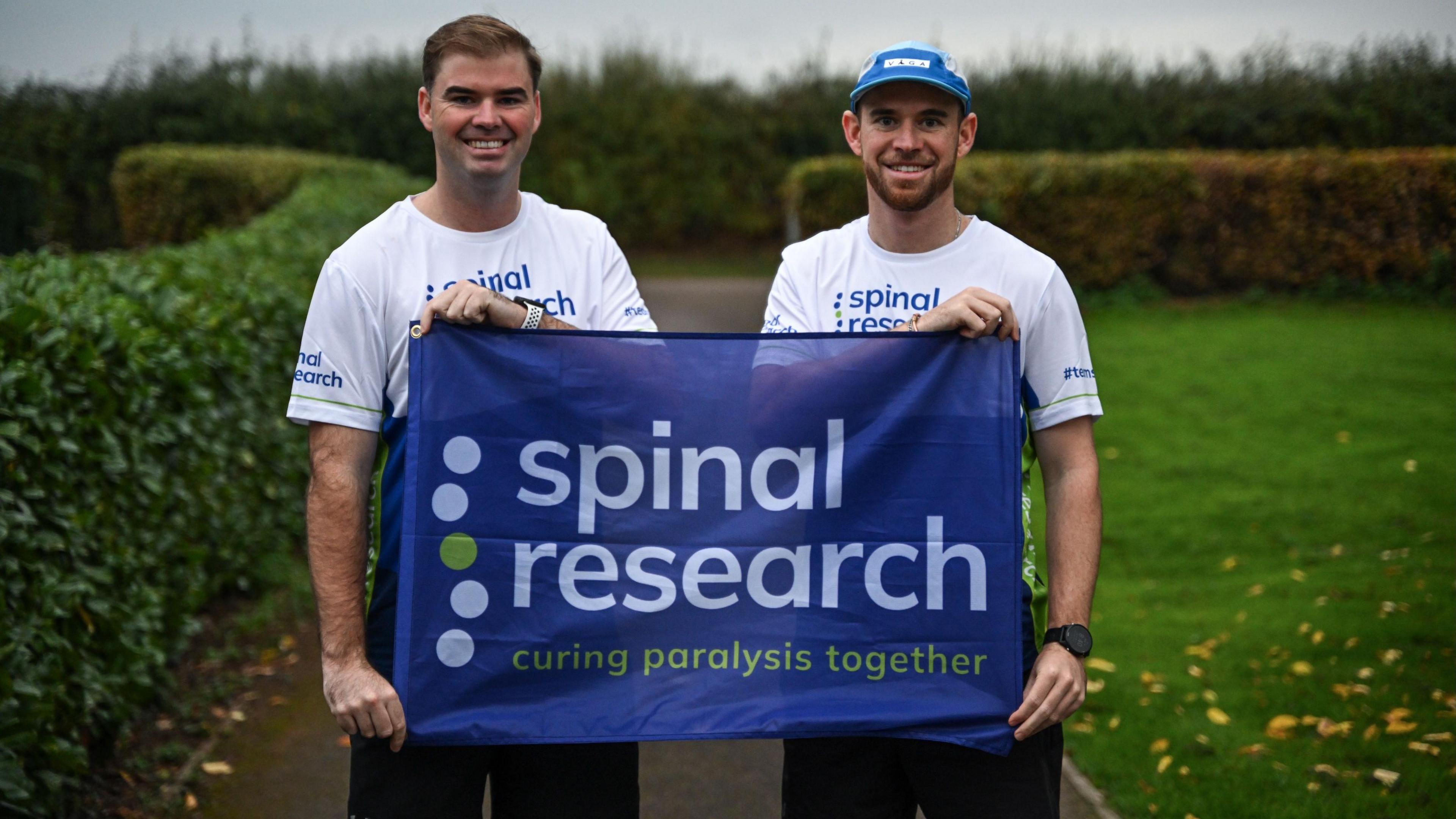 Two brothers in their late 20s or early 30s stand in a park lined with hedges, smiling and holding a blue banner which reads "Spinal research curing paralysis together". They are wearing white T-shirts with the Spinal Research logo on too, with the one on the right wearing a blue cap. There are yellow, fallen leaves strewn across the lawn.