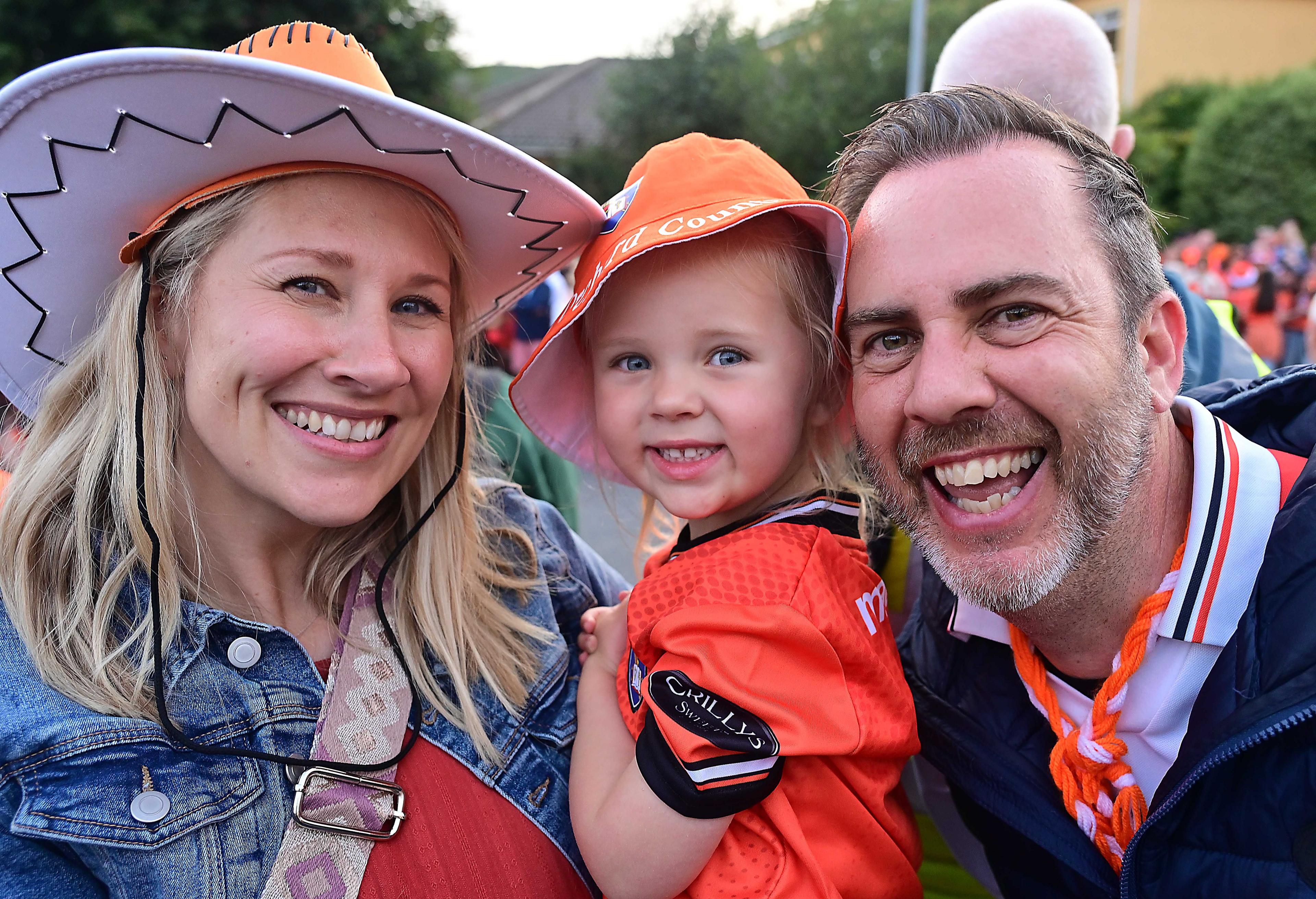 A man, woman and child. The child is in orange. The woman is wearing a cowboy hat.