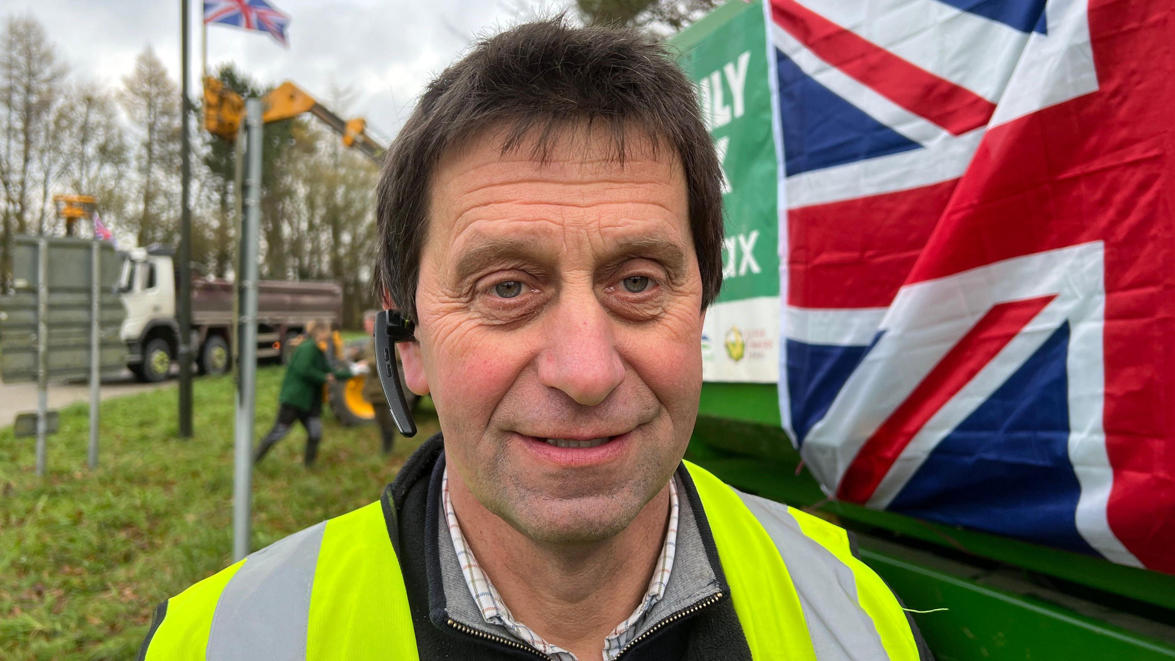 Brian Brasington wearing a yellow hi-vis vest and standing on a grassy verge beside a roundabout. Behind him there is a large green harvester with a Union Jack flag on the side of it, and several green tractors. 