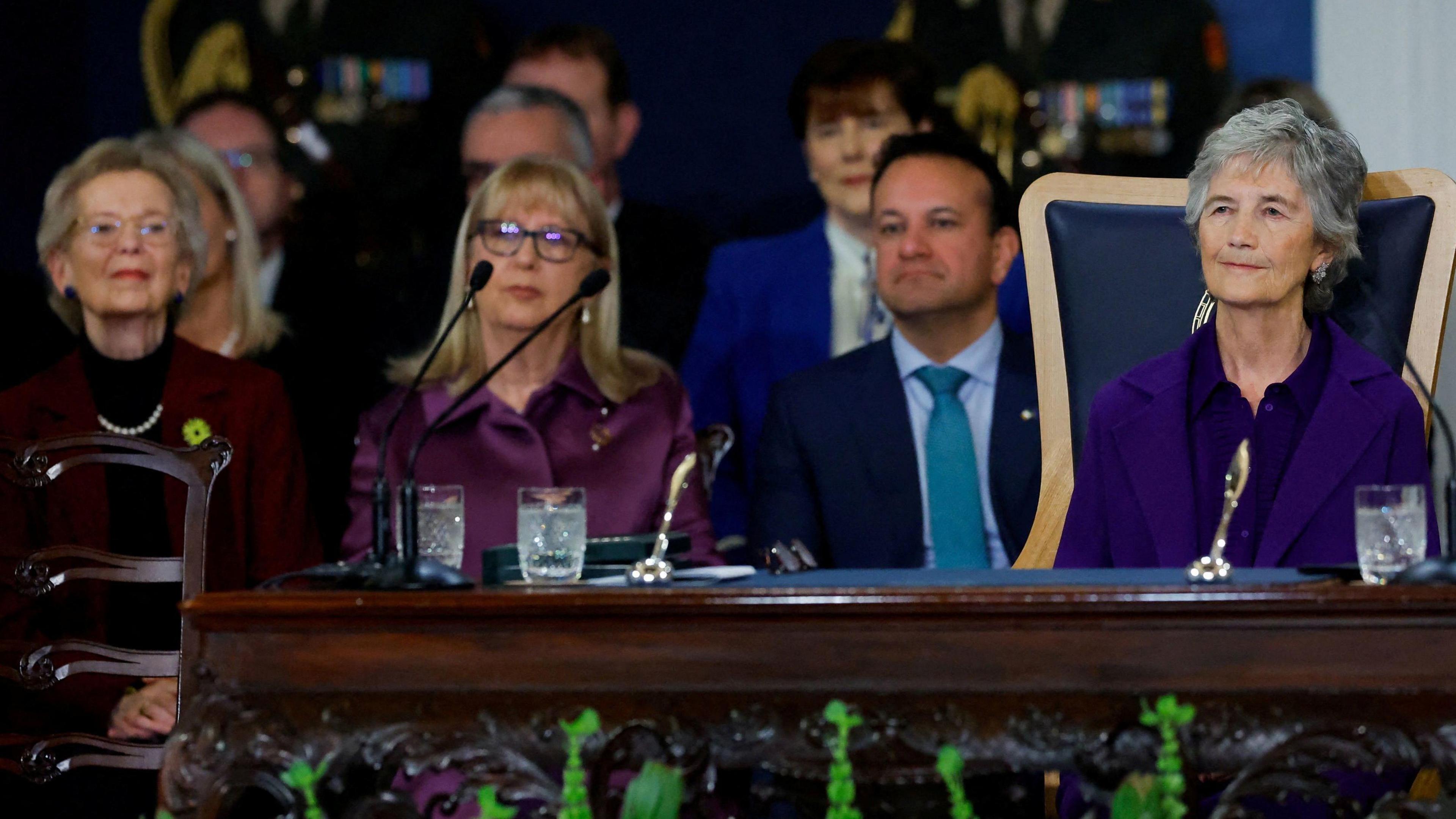 Catherine Connolly with short grey hair, wearing a purple top and purple jacket, sits at an ornate table. In the background are Mary Robinson, who has short grey hair and is wearing a purple jacket over a black jumper; Mary McAleese who has shoulder length blonde hair, glasses and is wearing a purple dress; and Leo Varadkar who has short black hair and wearing a dark blue jacket and a blue shirt and tie.