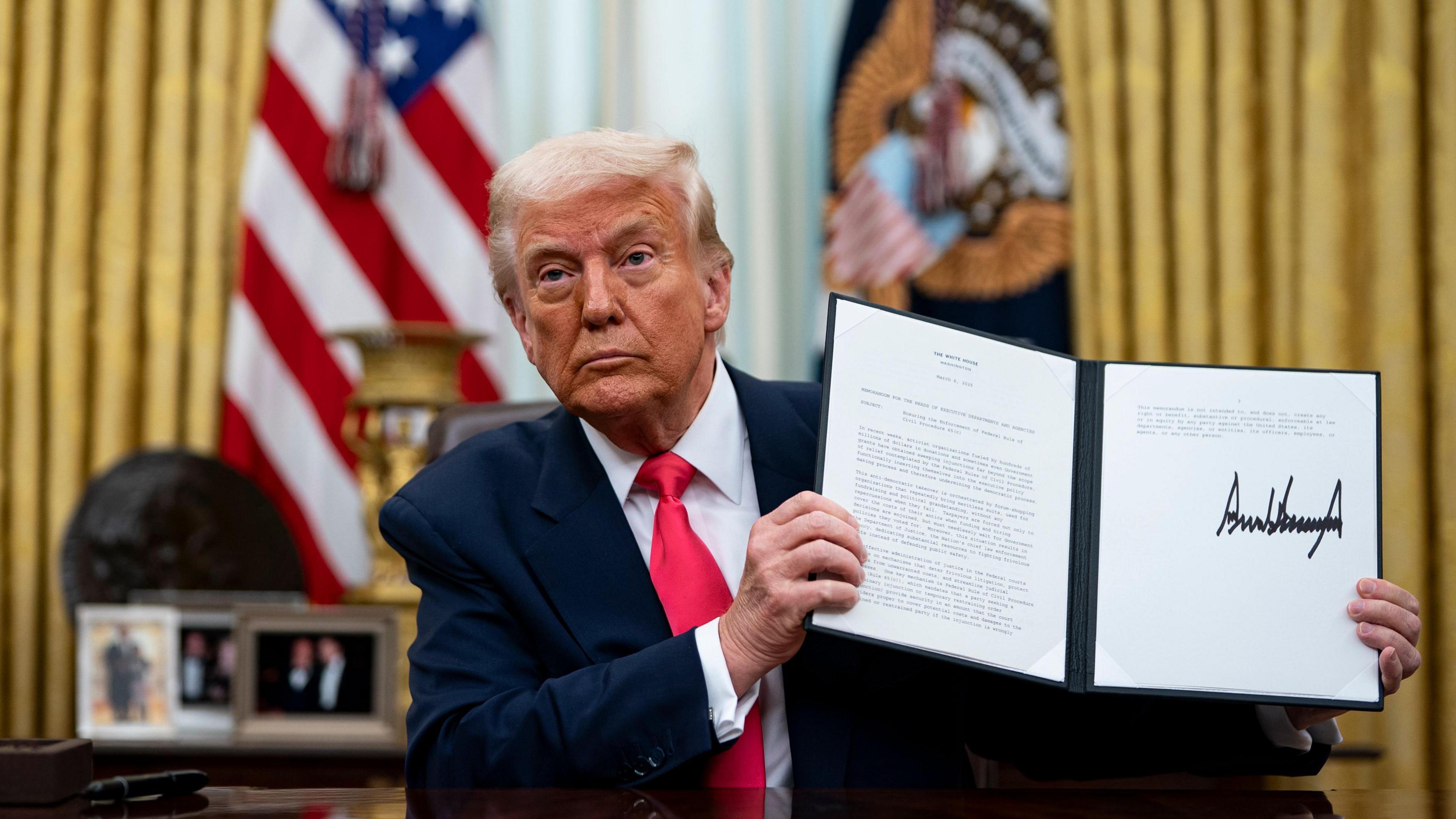 Donald Trump in the Oval Office holding up an executive order bearing his signature. He is wearing a blue suit and red tie. Behind him are the US flag, presidential flag, gold curtains and some photos.