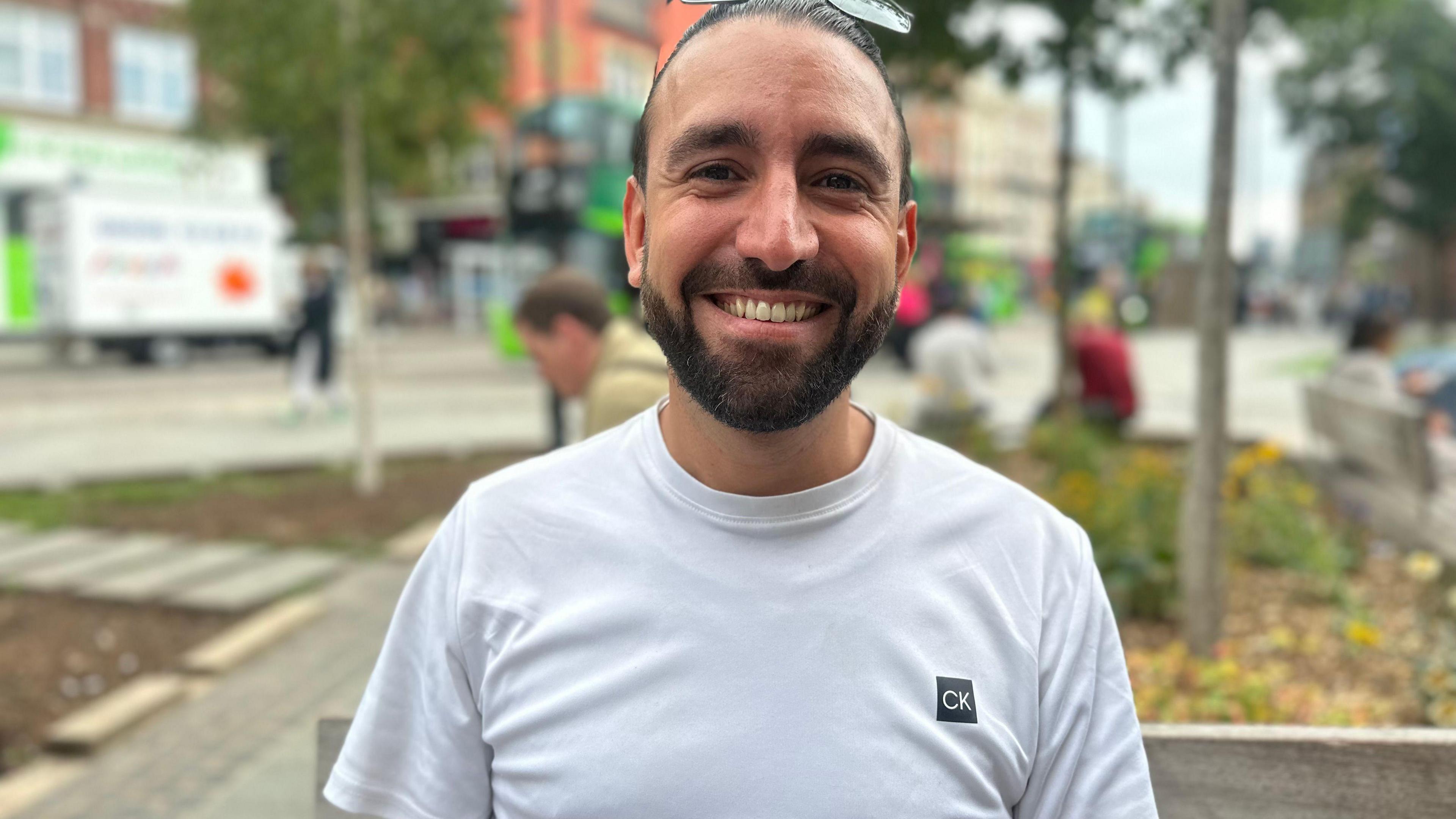 A young man with slicked back hair and glasses on his head and a neatly trimmed beard. He is wearing a white t-shirt and sat on a city centre bench.