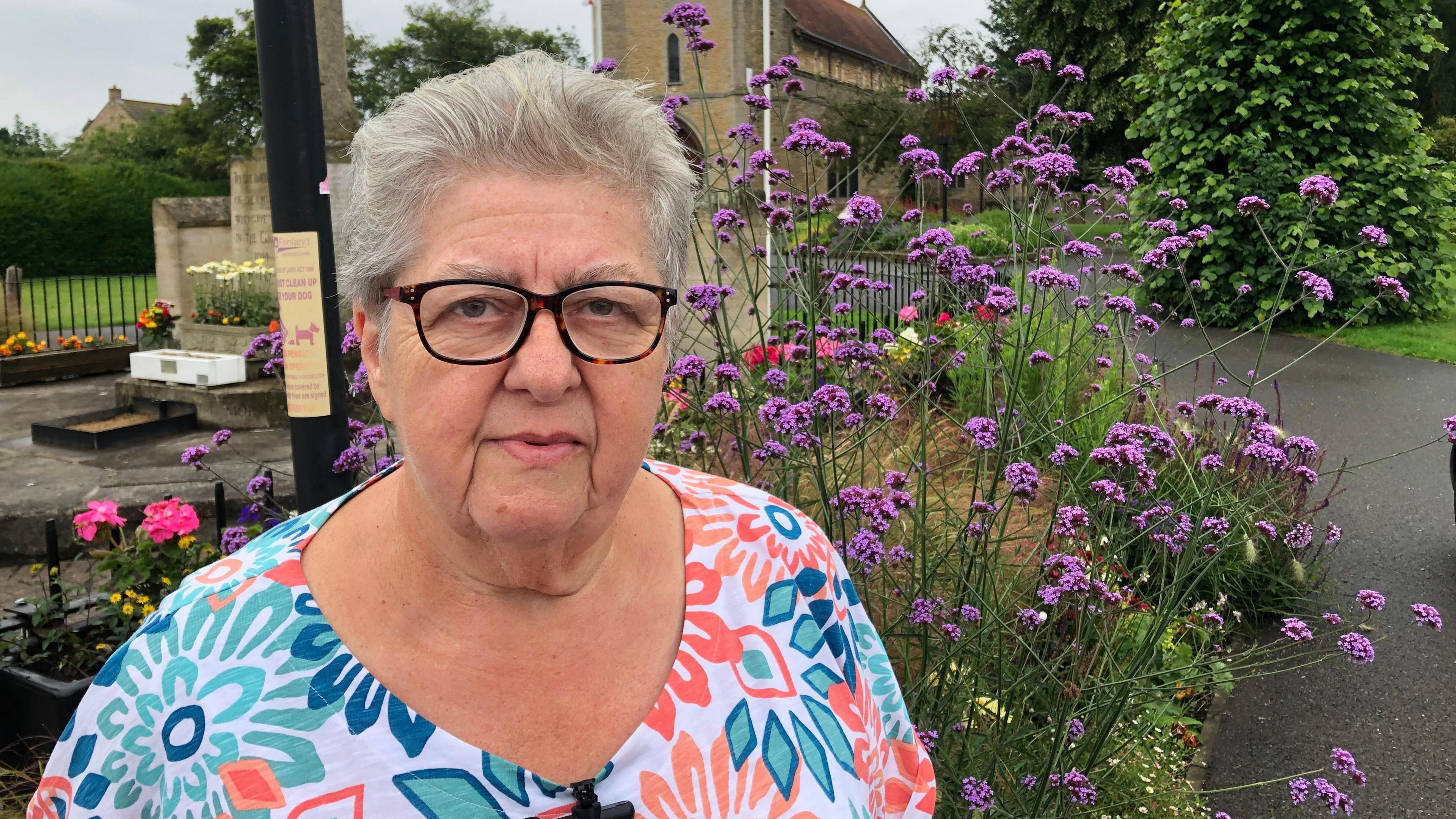 Anne Wells, chair of Chatteris in Bloom, outside the parish church in town. She is wearing a white top covered in multi-coloured flowers.