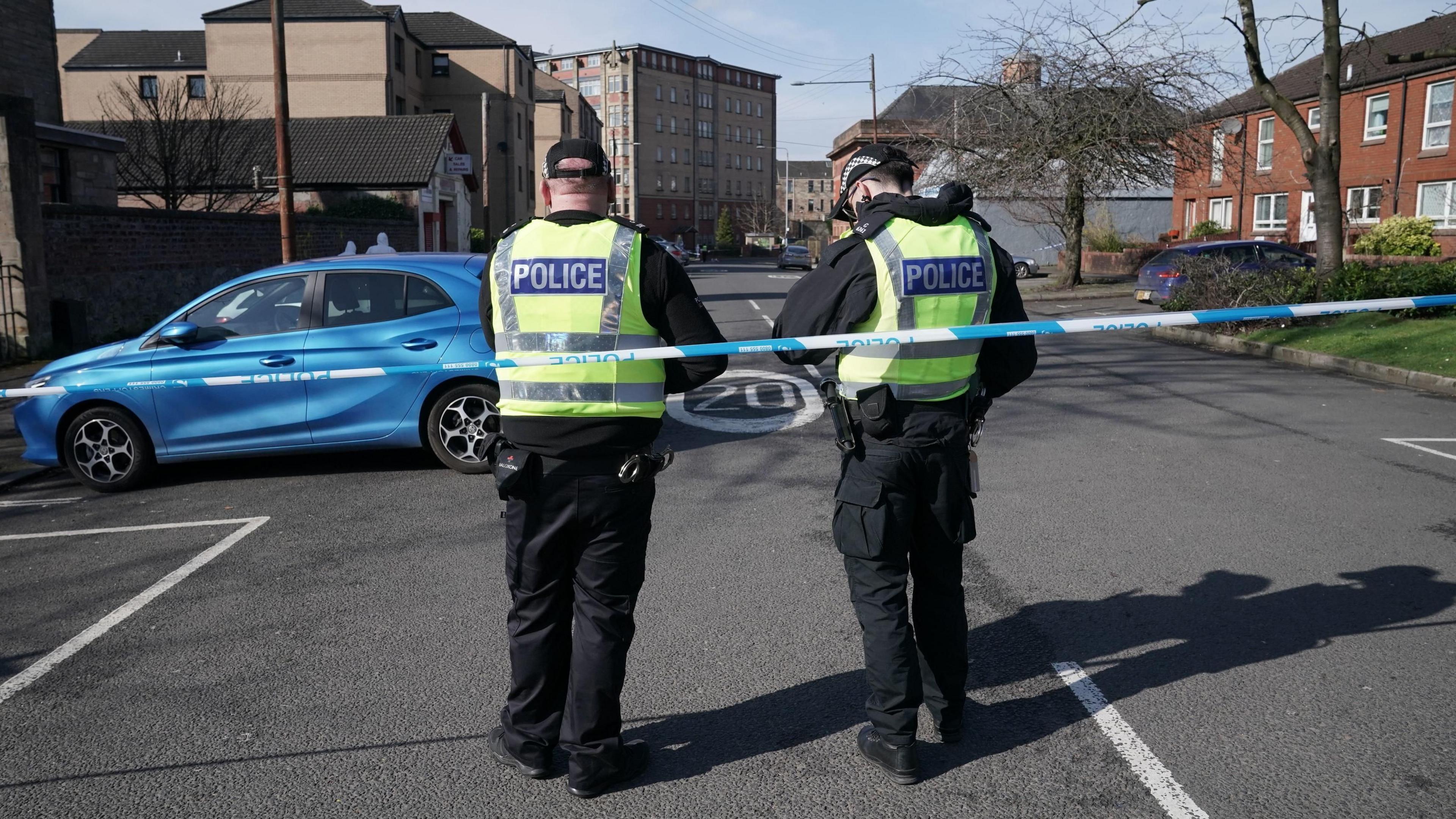 Two constabulary  officers basal   down  constabulary  tape, with their backs to the camera. Flats and houses tin  beryllium  seen successful  the background, arsenic  good   arsenic  a bluish  car.