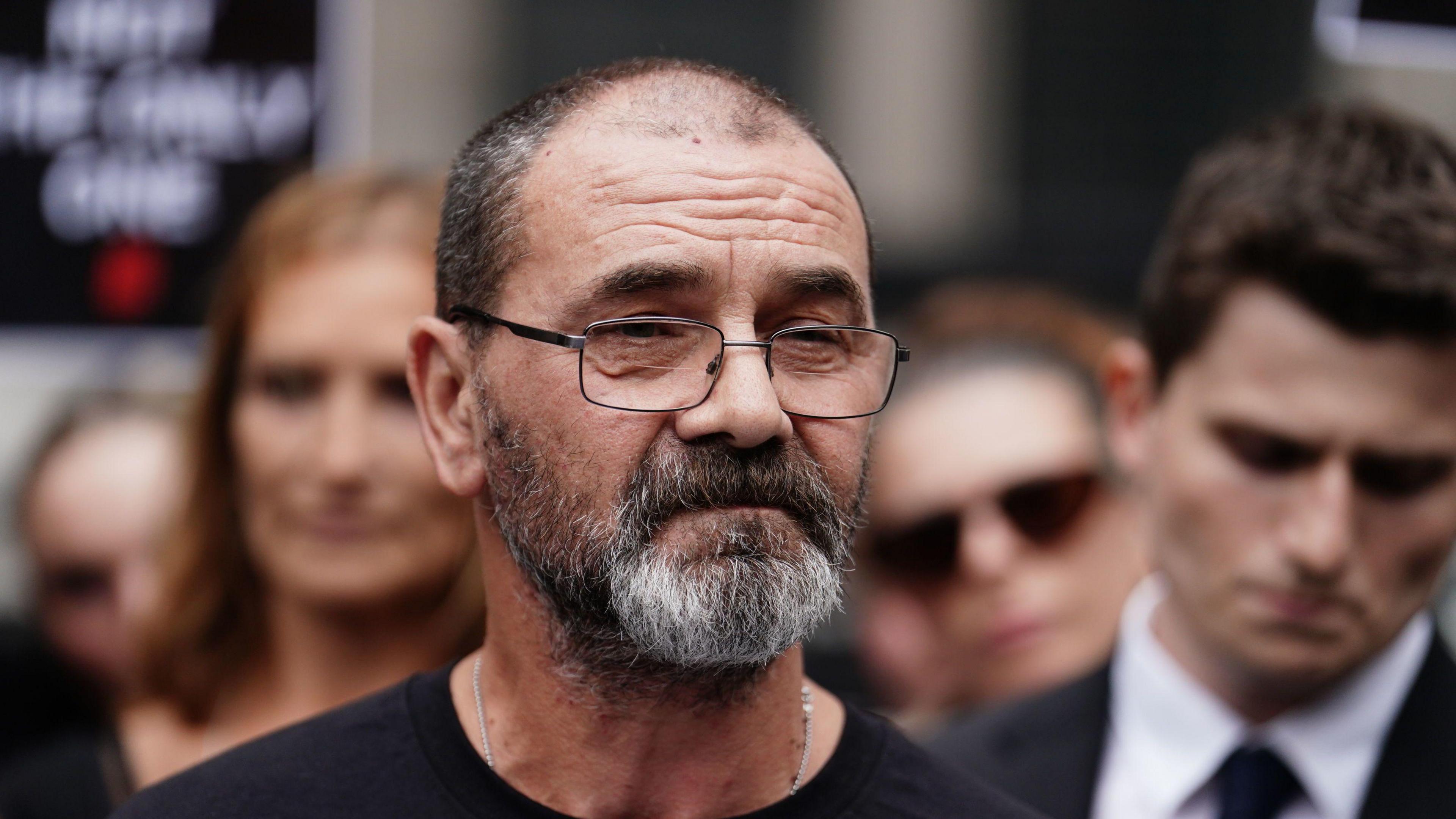 Andrew Malkinson wears glasses and has a greying bead as he looks solemn outside court 