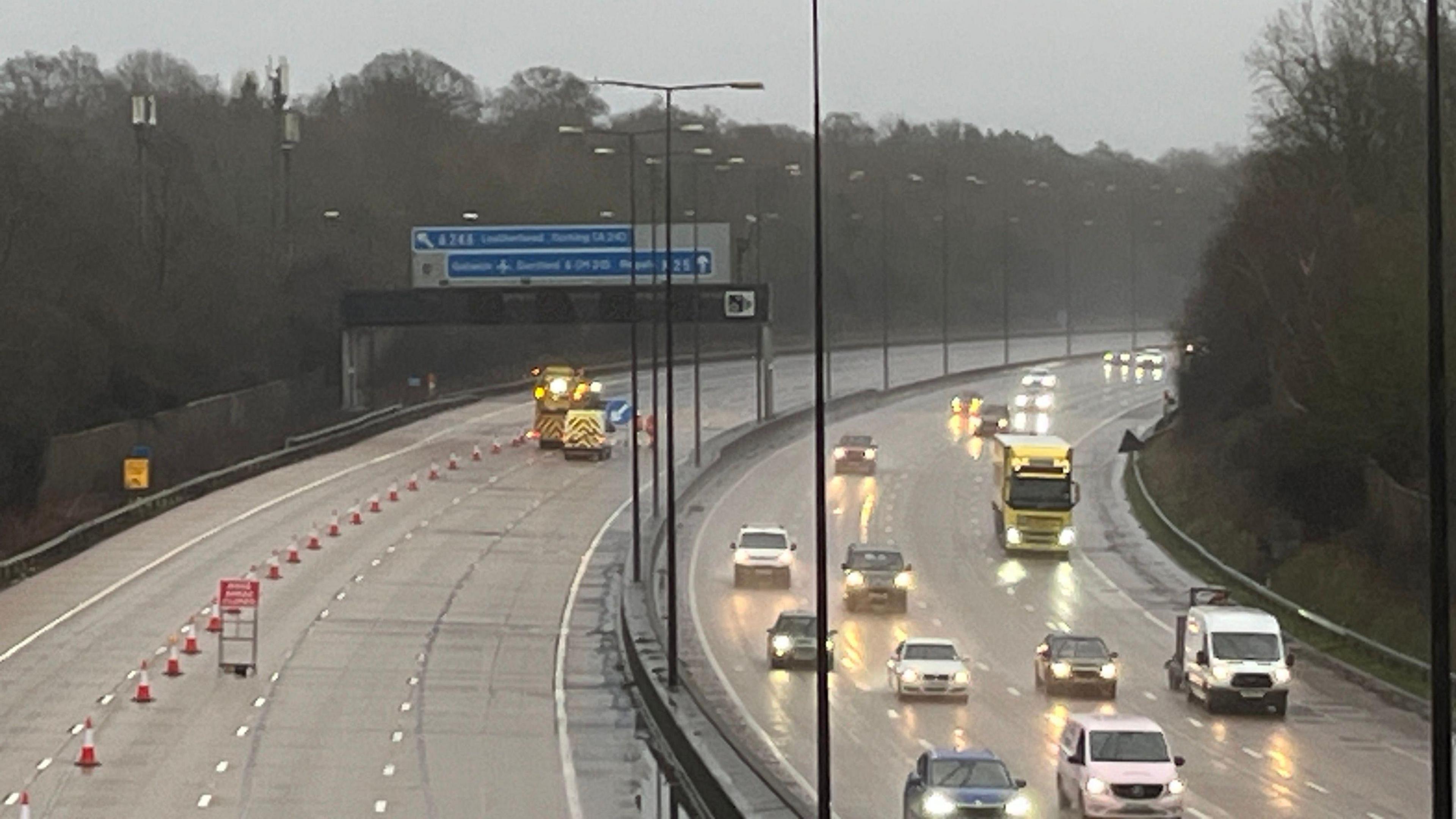 The anti-clockwise carriageway of the M25 from Oaklawn Rd bridge over M25 in Leatherhead, closed to traffic, with cones having been put out. Traffic is still moving on the clockwise carriageway, on a grey and wet day.