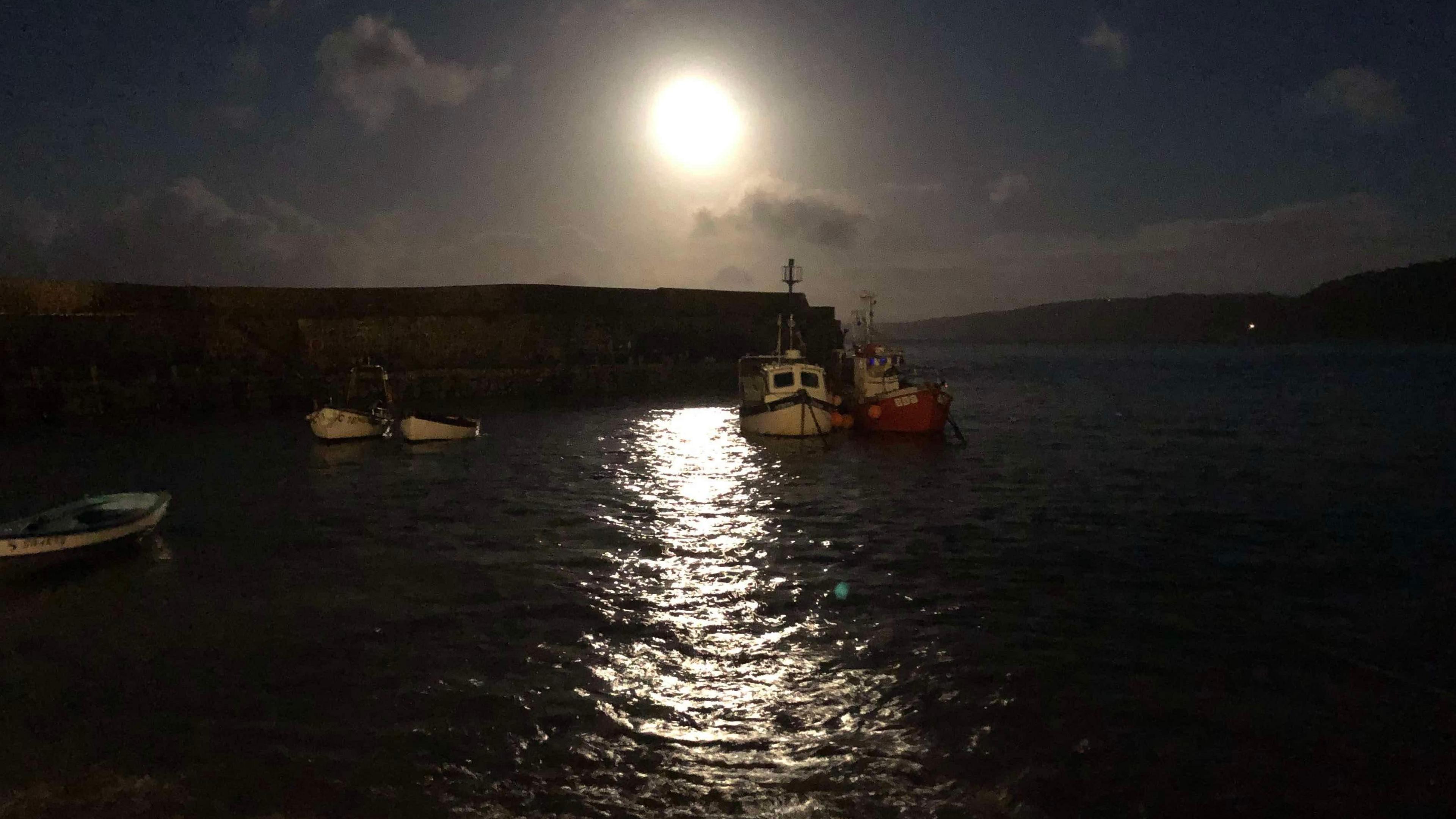 The full moon shines over a harbour, with boats in the foreground and the moonlight reflecting off the water