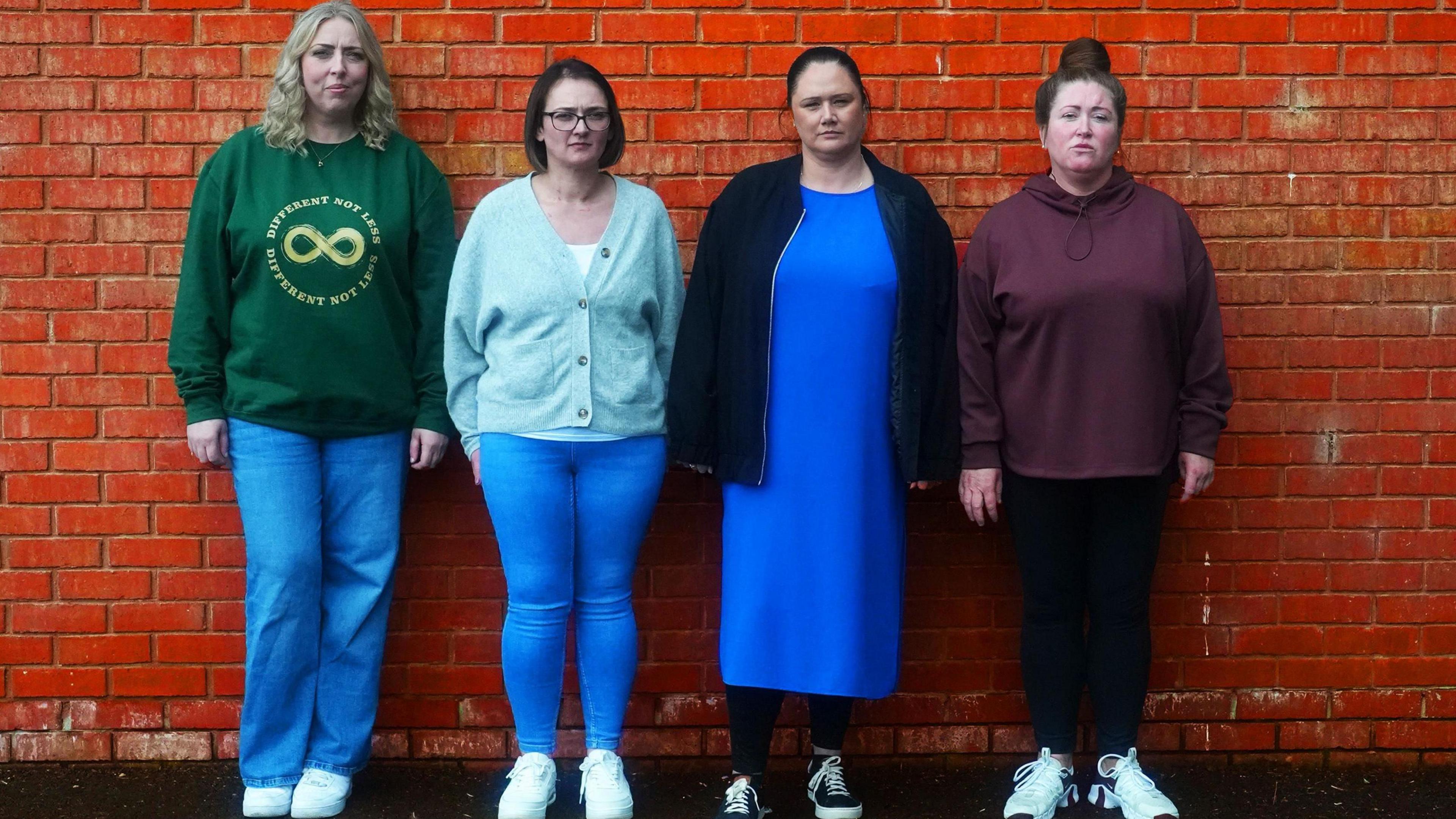 Four women. Far left has blonde hair and is wearing a green sweater and blue jeans, next to her has shot brown hair and is wearing a sky blue cardigan and blue jeans, next to her has brown hair pulled back into a pony tail, a blue dress and darker blue jacket and next to her has brown hair in a top knot, a maroon hoody and dark blue leggings
